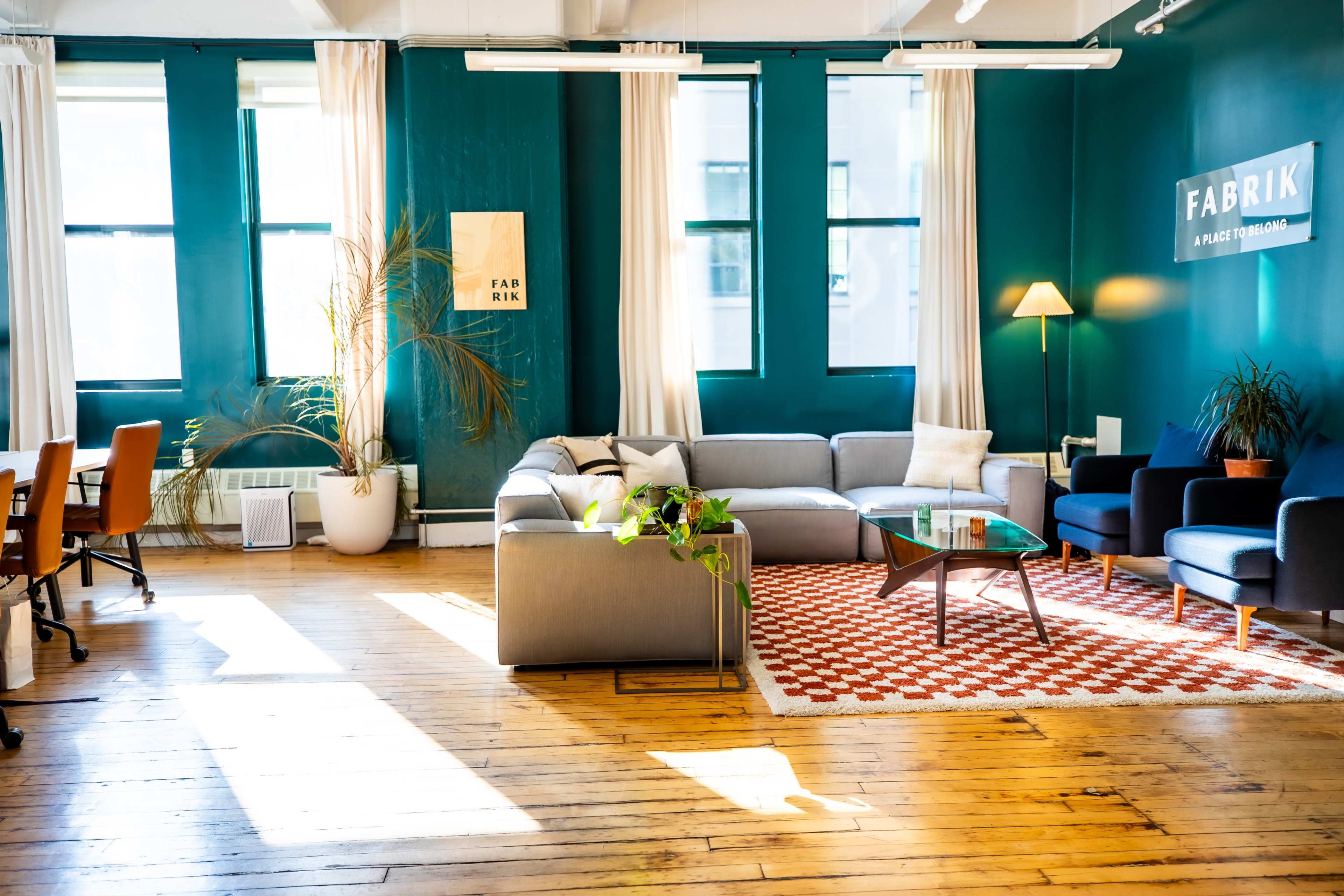 The image shows a modern, bright co-working space with teal walls, comfortable seating, and large windows allowing natural light to fill the room.