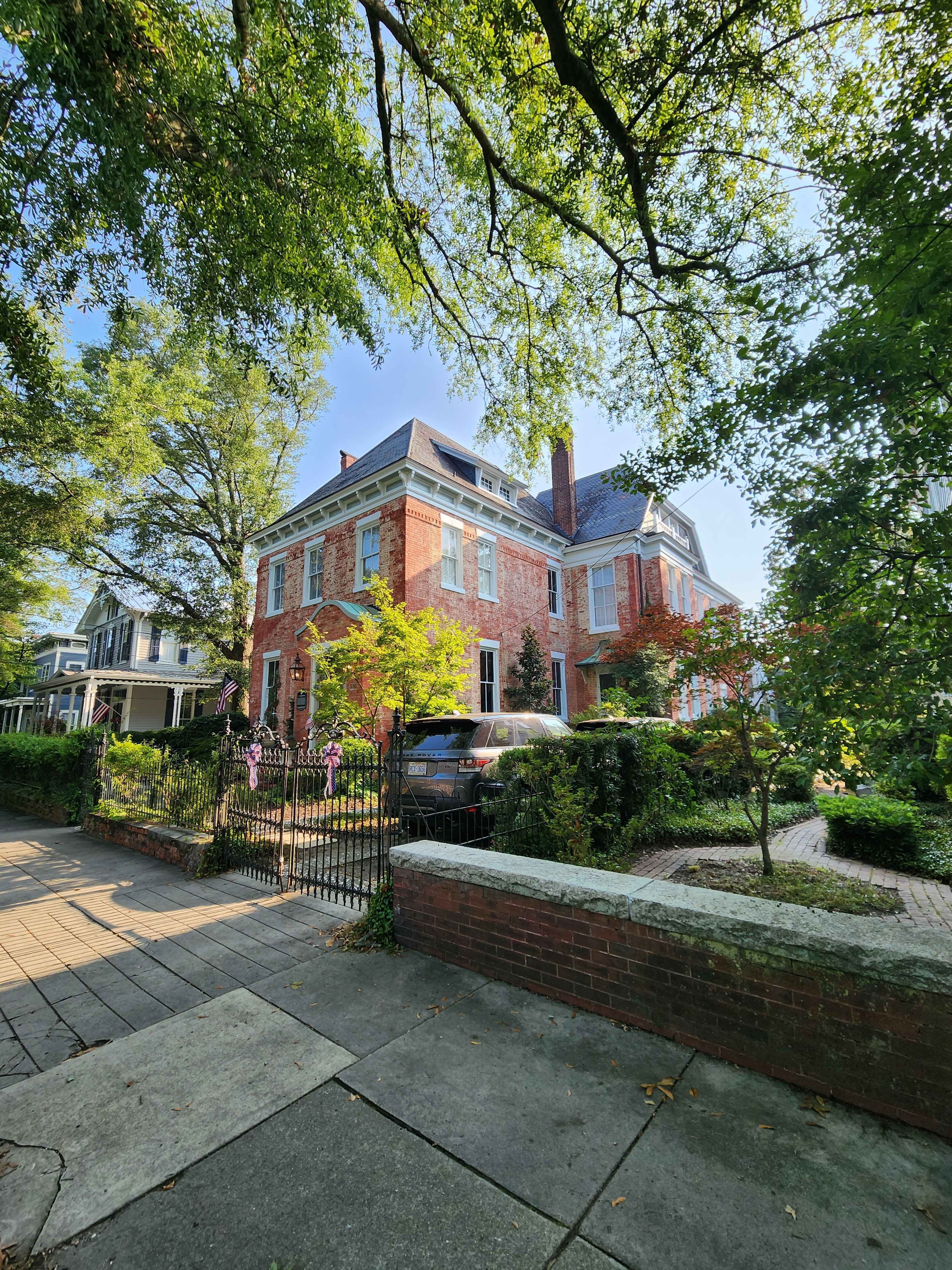 A large red brick house with a steep roof is surrounded by greenery and a wrought iron fence.