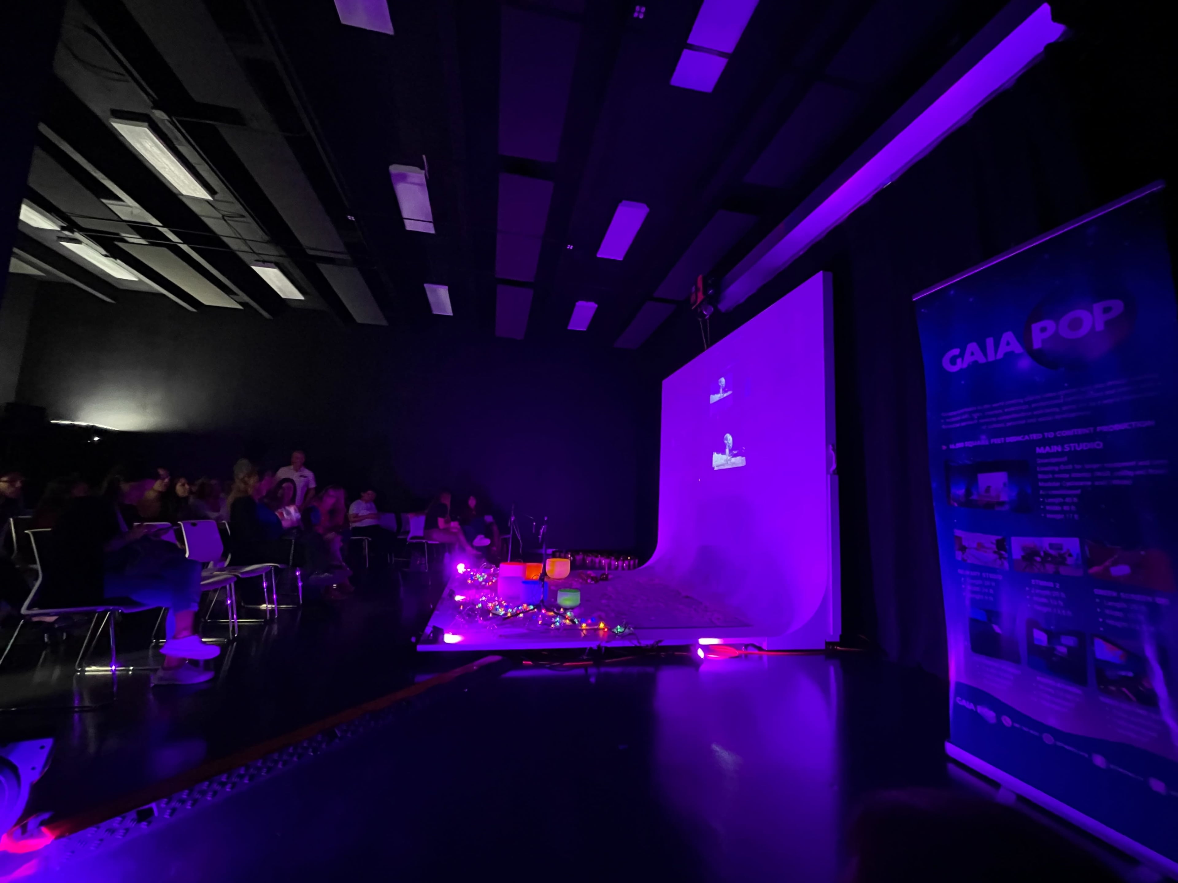 A stage is set with a purple backdrop and several colorful objects arranged in front of an audience in a dimly lit room.