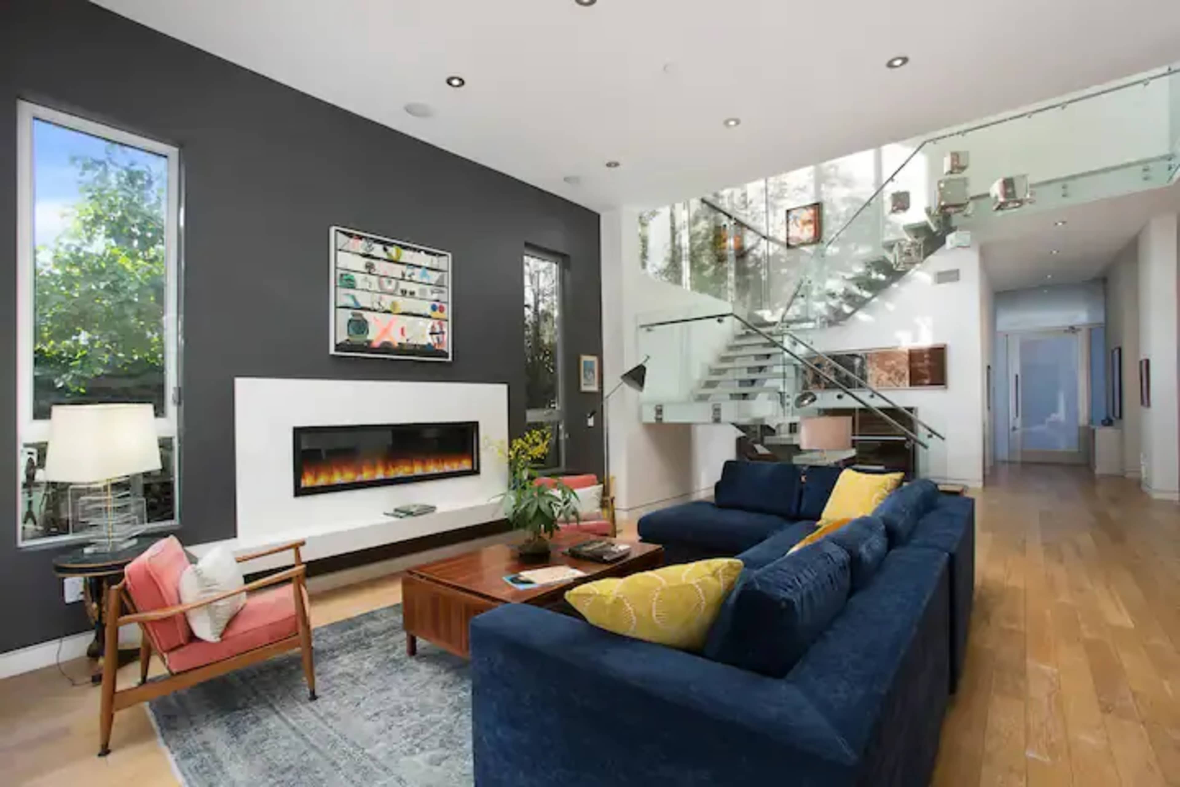 A modern living room with a large blue sofa, a wooden coffee table, and a wall-mounted fireplace, featuring tall windows and a staircase with glass railings.