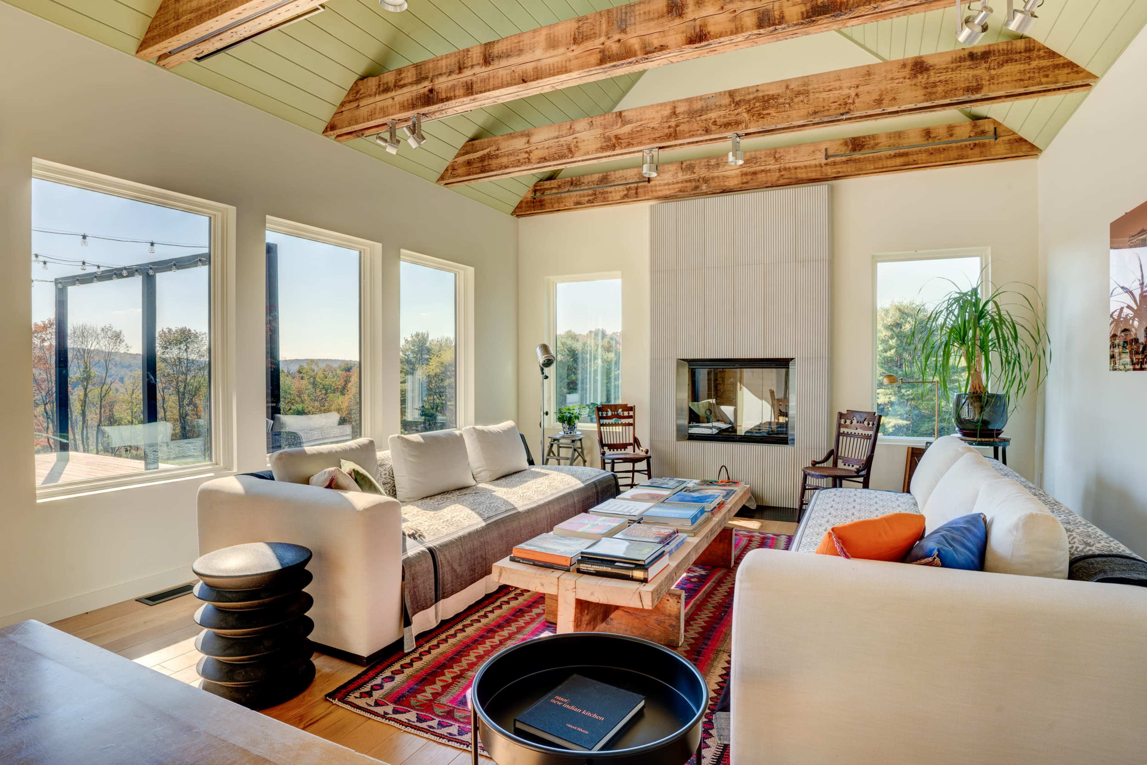 A spacious living room features large windows, exposed wooden beams, and a central fireplace, with comfortable furniture and books spread across a coffee table.