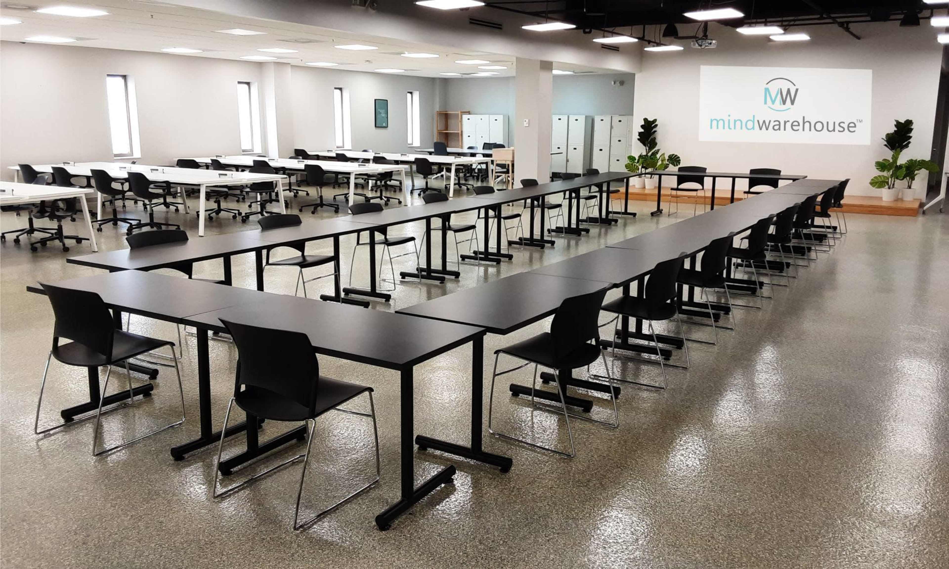A spacious training room with multiple rows of tables and chairs arranged neatly, along with a branded wall in the background.