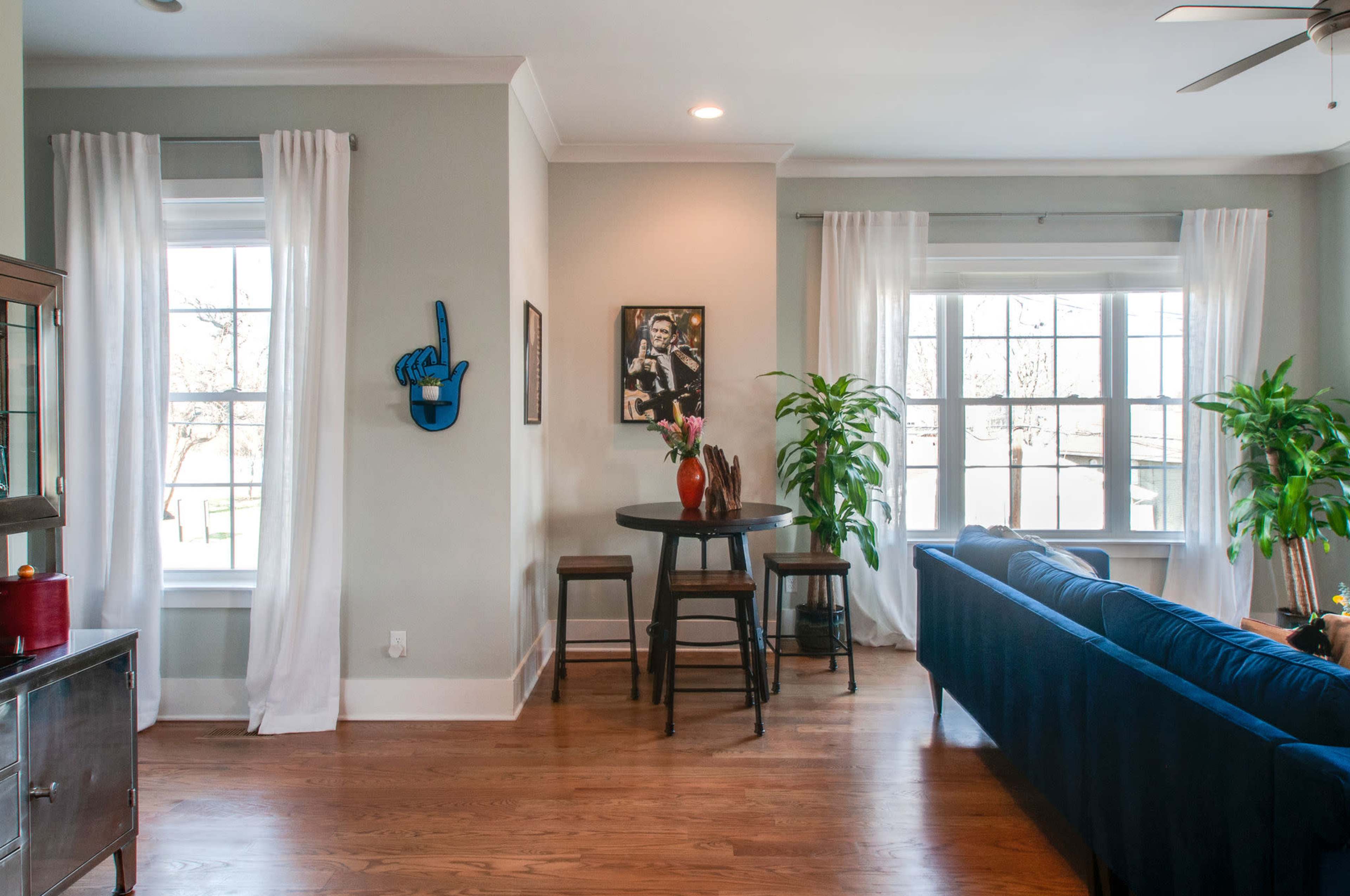 A living room features a round table with chairs, a blue couch, potted plants, and large windows adorned with white curtains.
