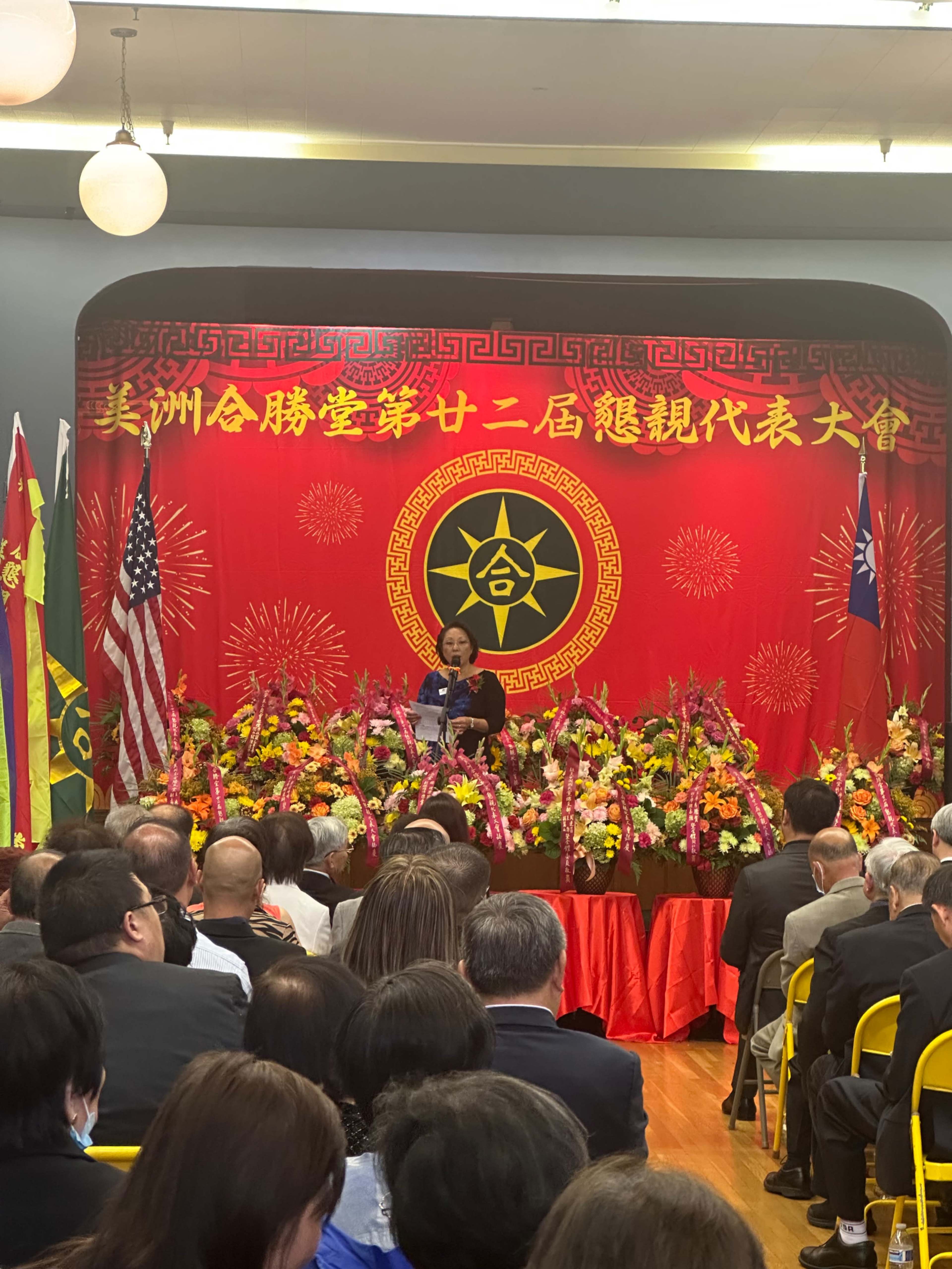 A speaker addresses an audience at a formal event decorated with vibrant banners and floral arrangements.