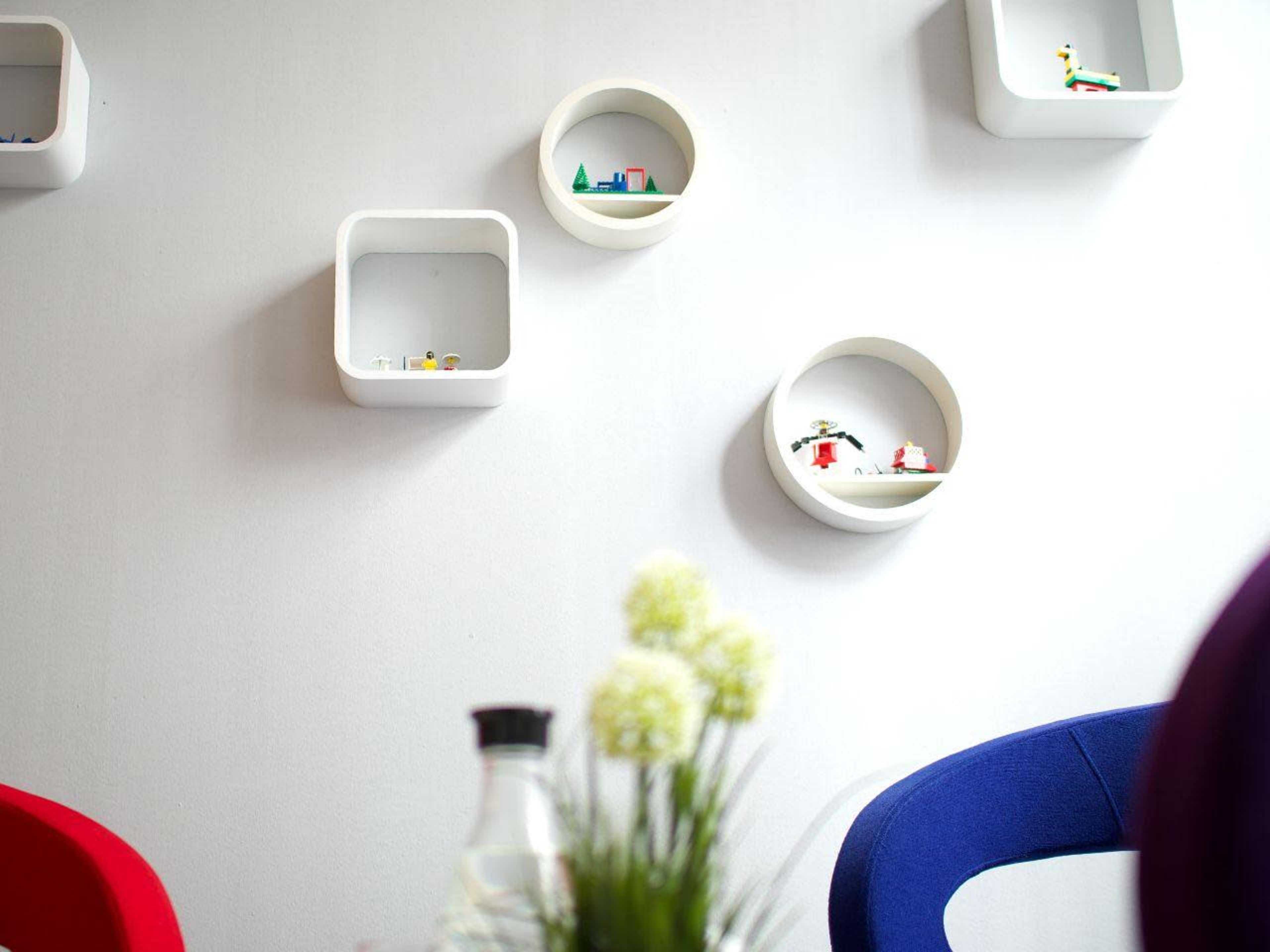 A minimalist interior with white wall-mounted shelves displaying small decorative items, and colorful chairs in the foreground.