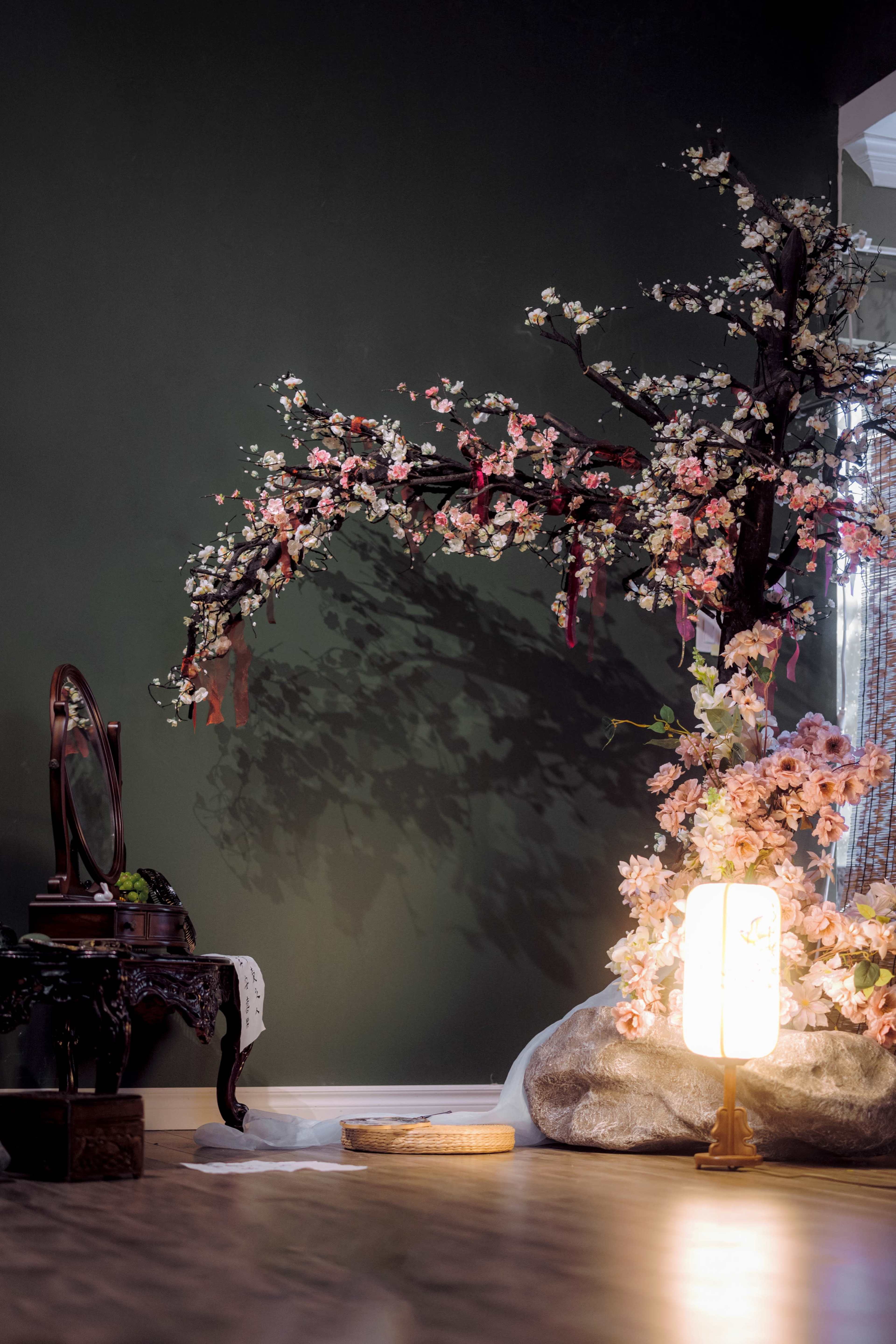A decorative display features a cherry blossom tree, a vintage mirror on a stand, and a lantern beside an arrangement of flowers on a dark green wall.