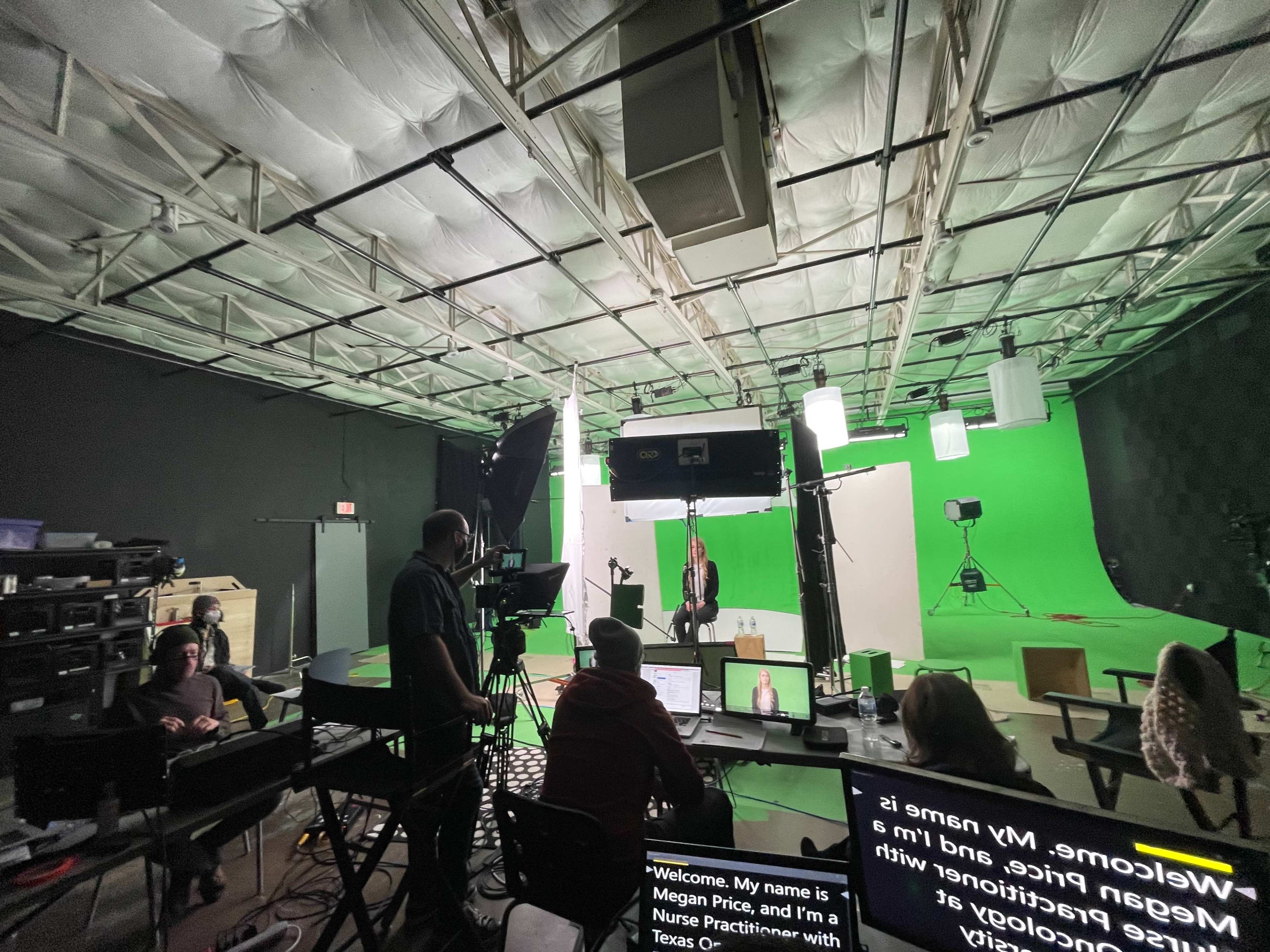 A professional video recording studio features a speaker in front of a green screen, with equipment and crew members visible in the background.