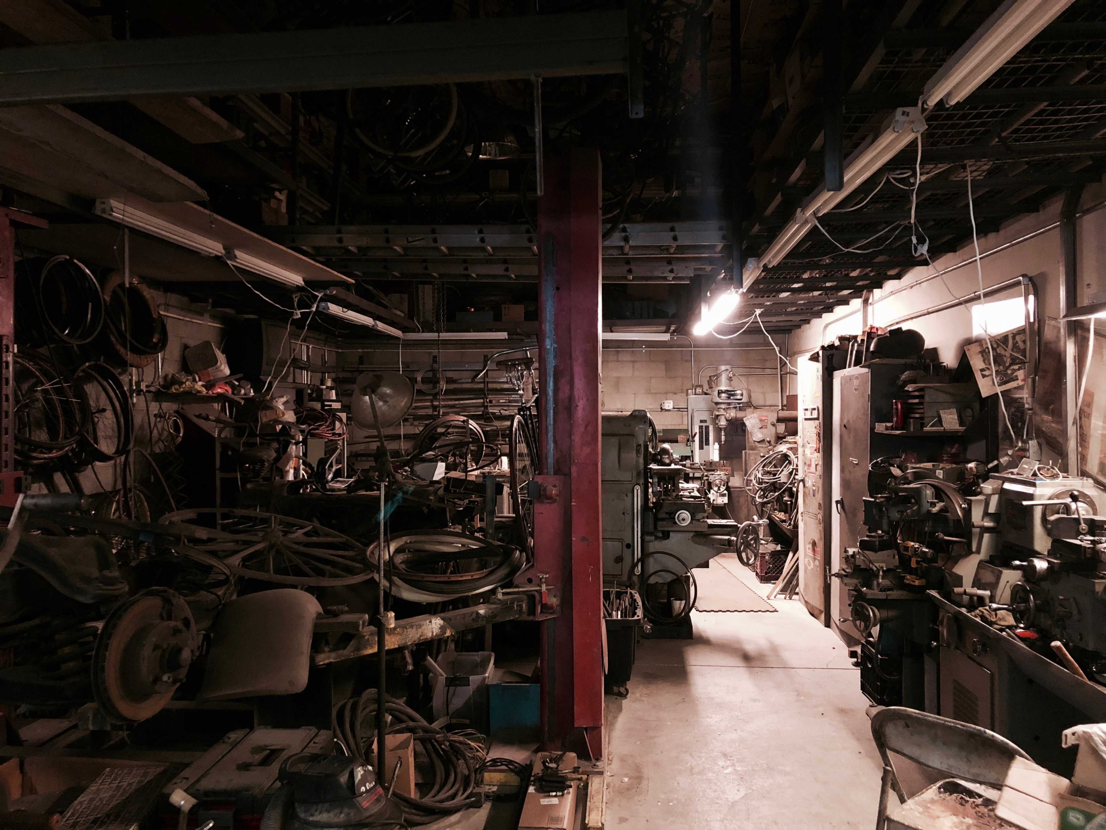 A cluttered workshop filled with various tools, machinery, and remnants of bicycles in dim lighting.