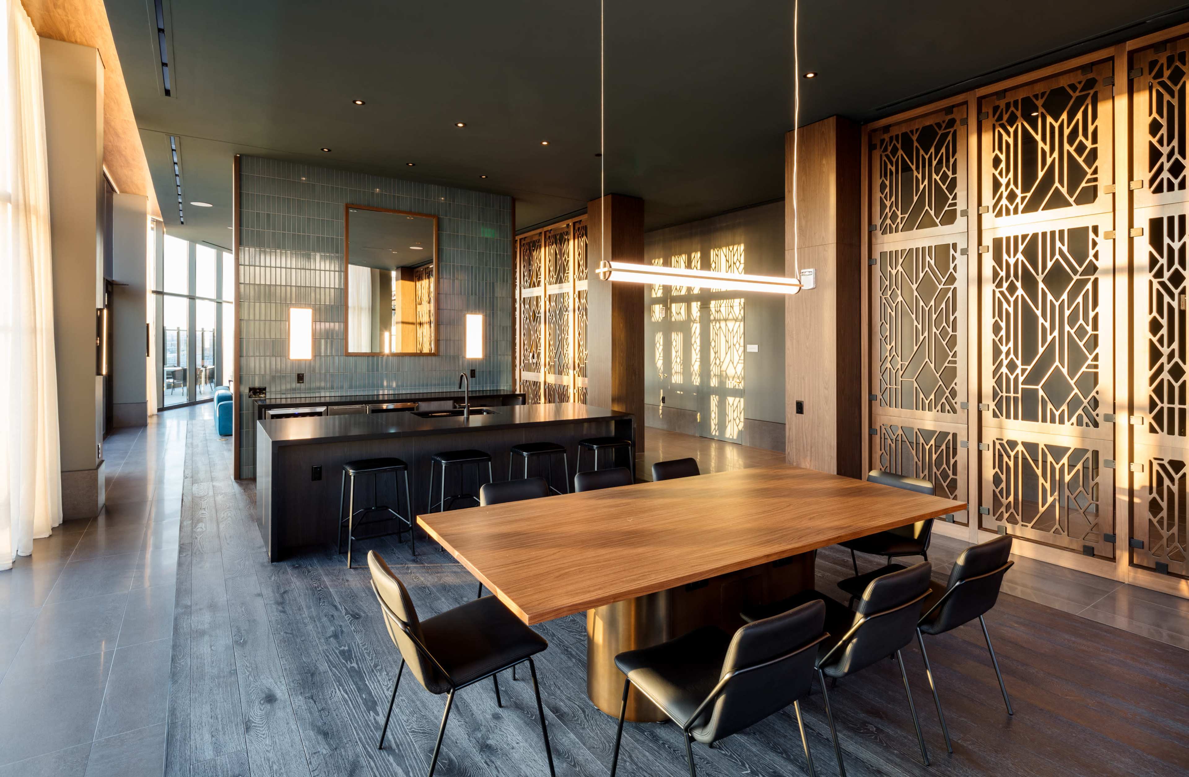 The image shows a modern kitchen and dining area featuring a large wooden table with black chairs, sleek cabinetry, and decorative wall screens.