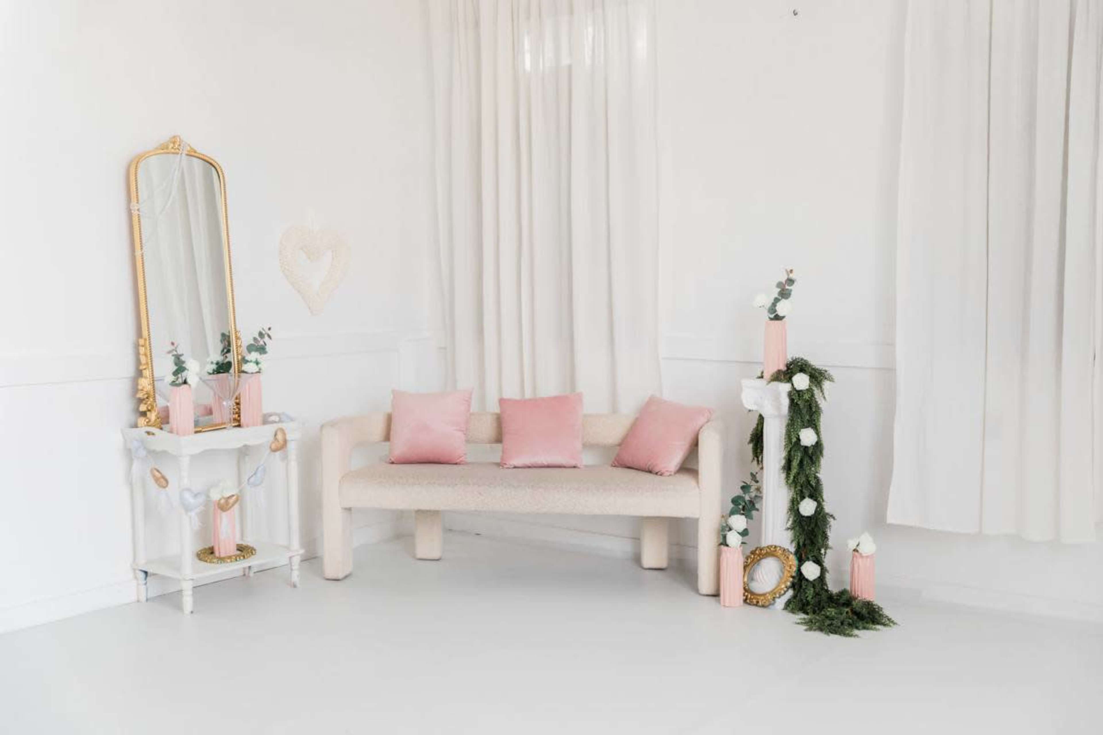 The image shows a minimalistic, light-colored interior with a sofa, decorative pillows, a mirror, and greenery accents.
