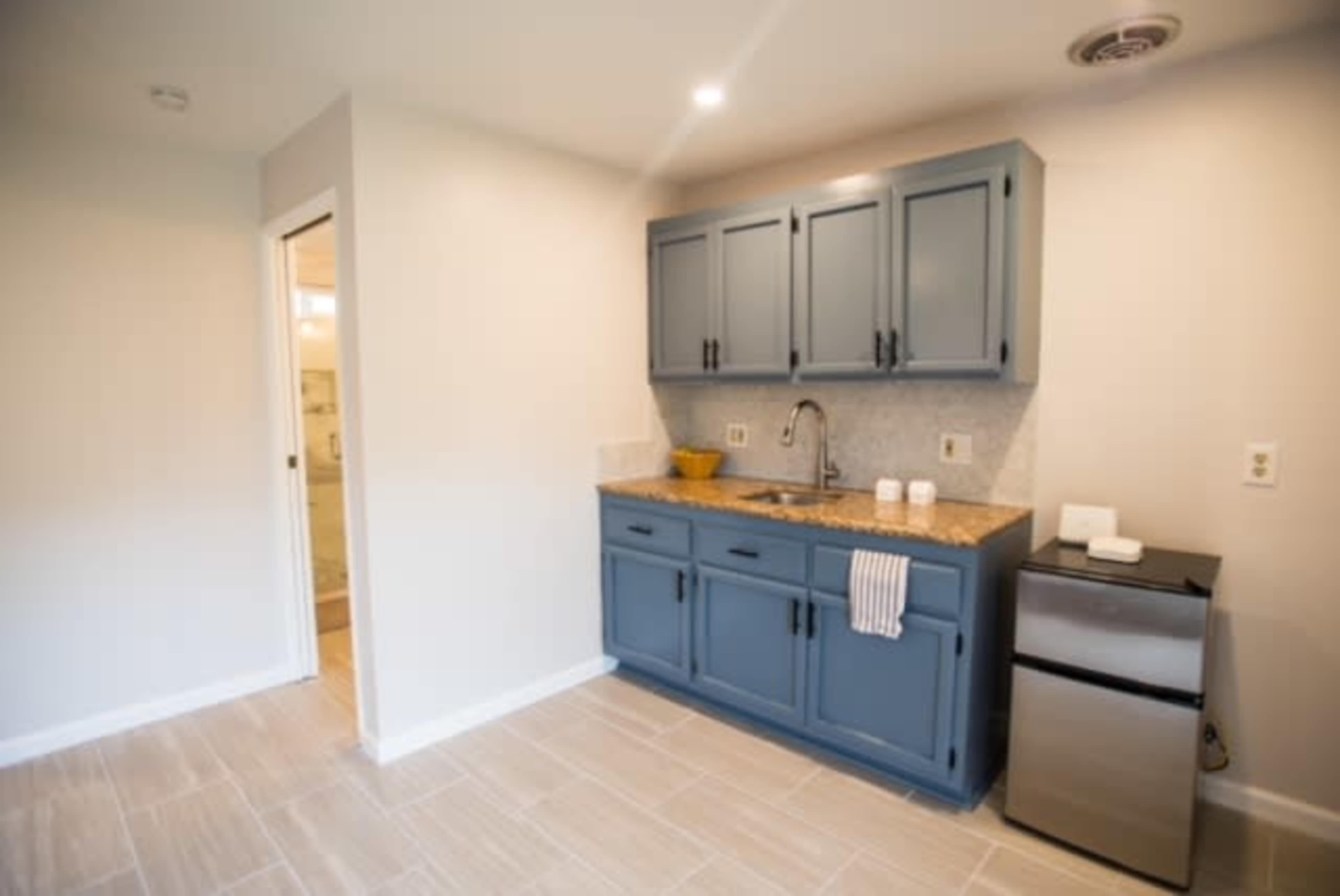 The image shows a compact kitchen area with blue cabinets, a granite countertop, a sink, and a small refrigerator, set against a light-colored wall and tiled floor.