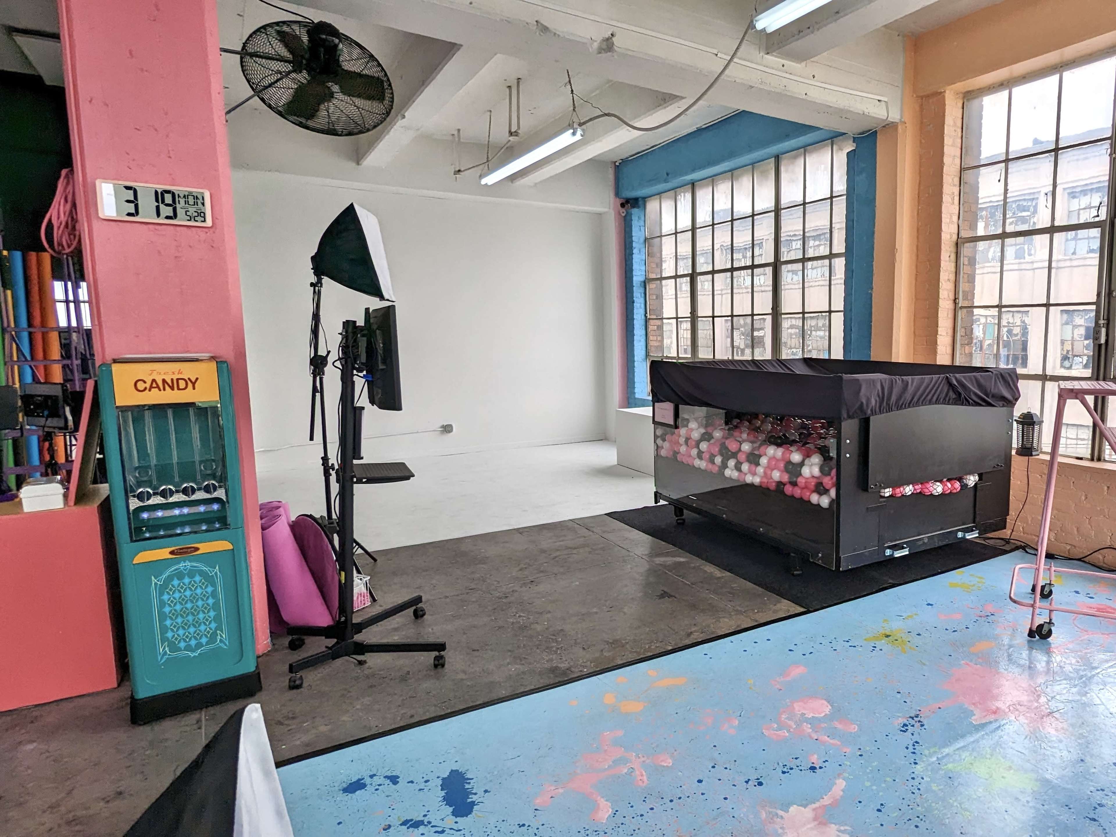 The image shows an empty studio space featuring a colorful floor, a candy vending machine, a large black display unit filled with balls, and large windows providing ample natural light.