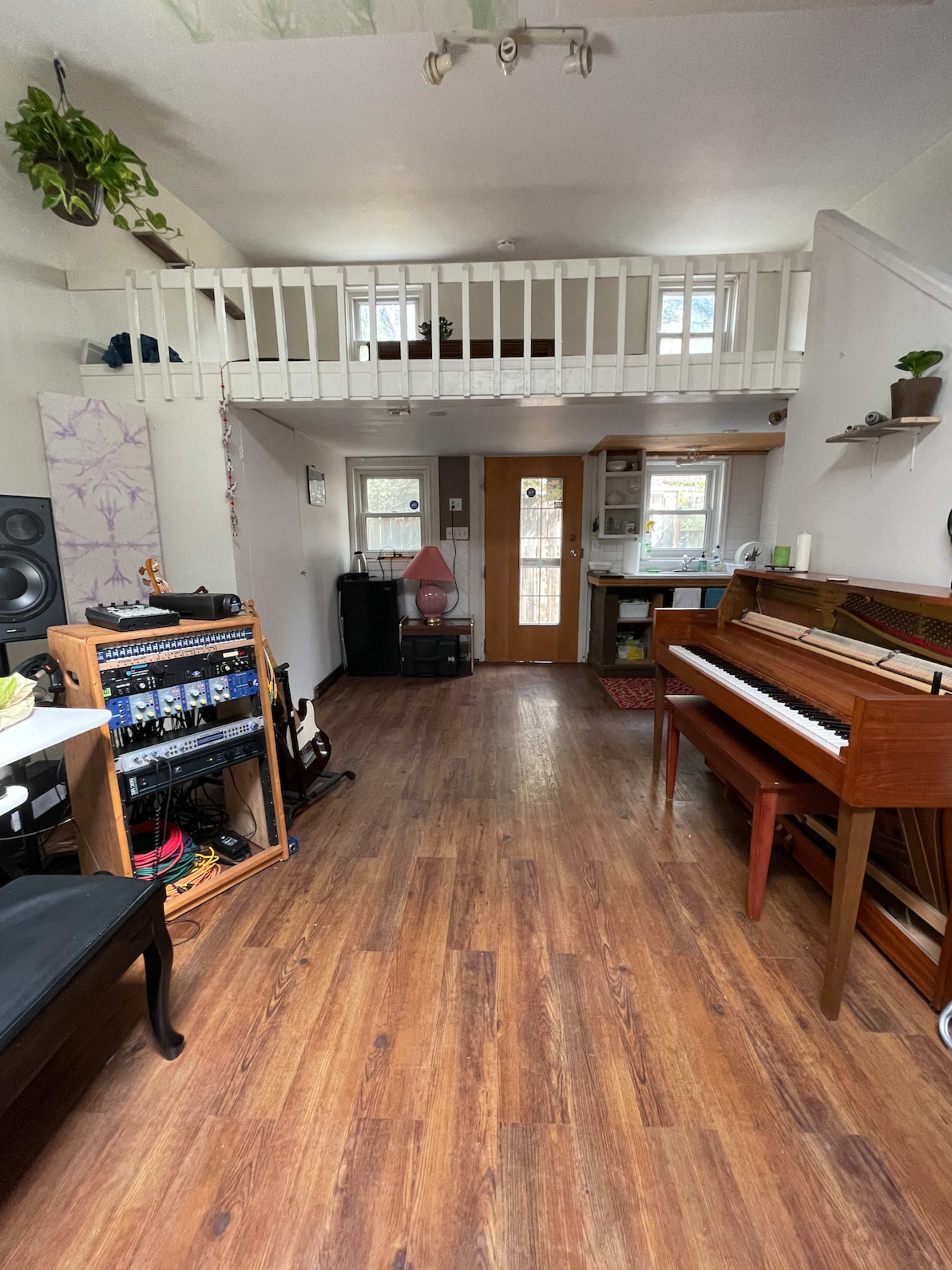 A spacious room with wooden flooring, including a piano, a sound equipment rack, and a loft area above.