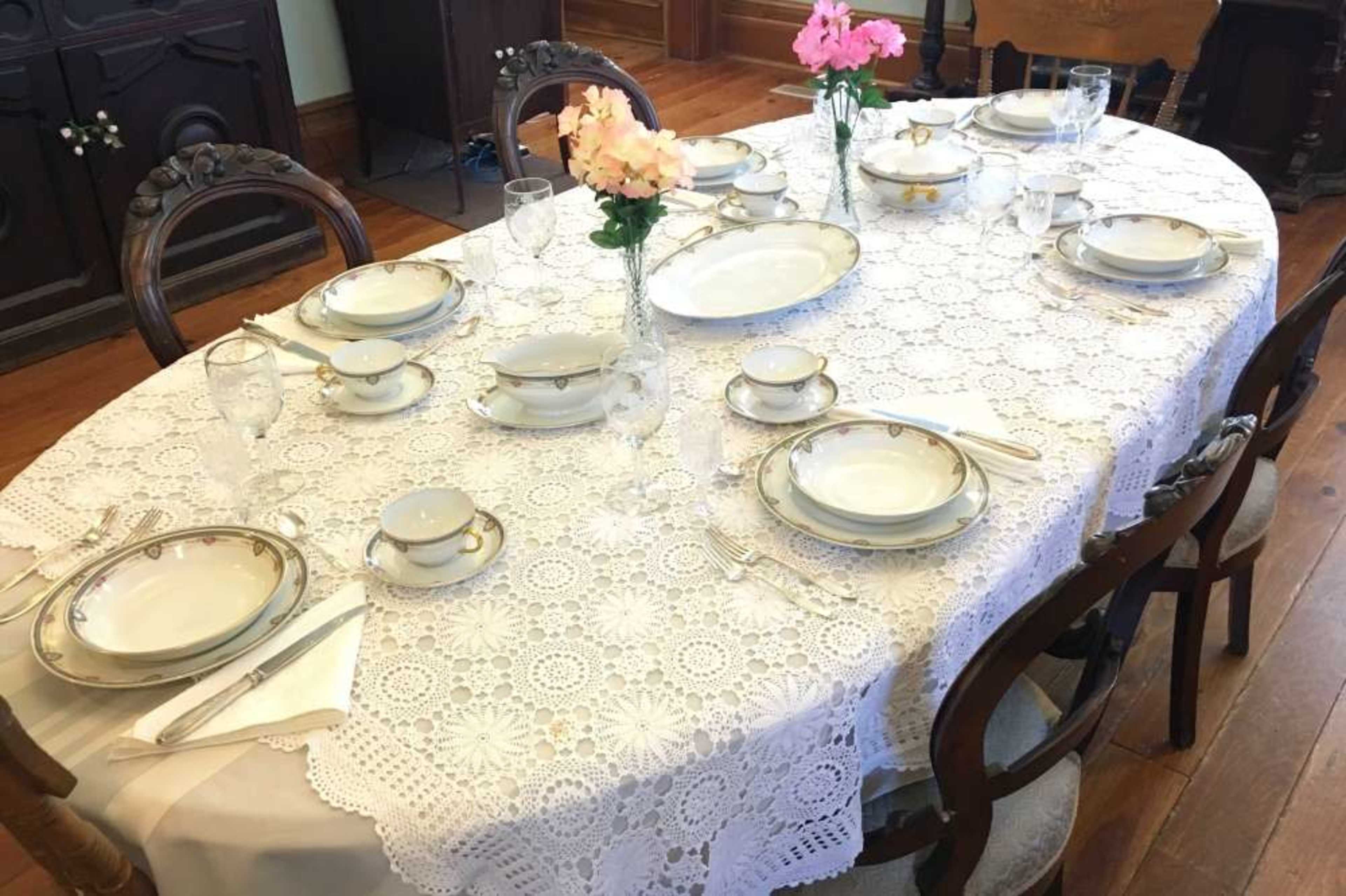 A dining table is set with elegant plates, cutlery, and floral centerpieces, covered by a lace tablecloth.