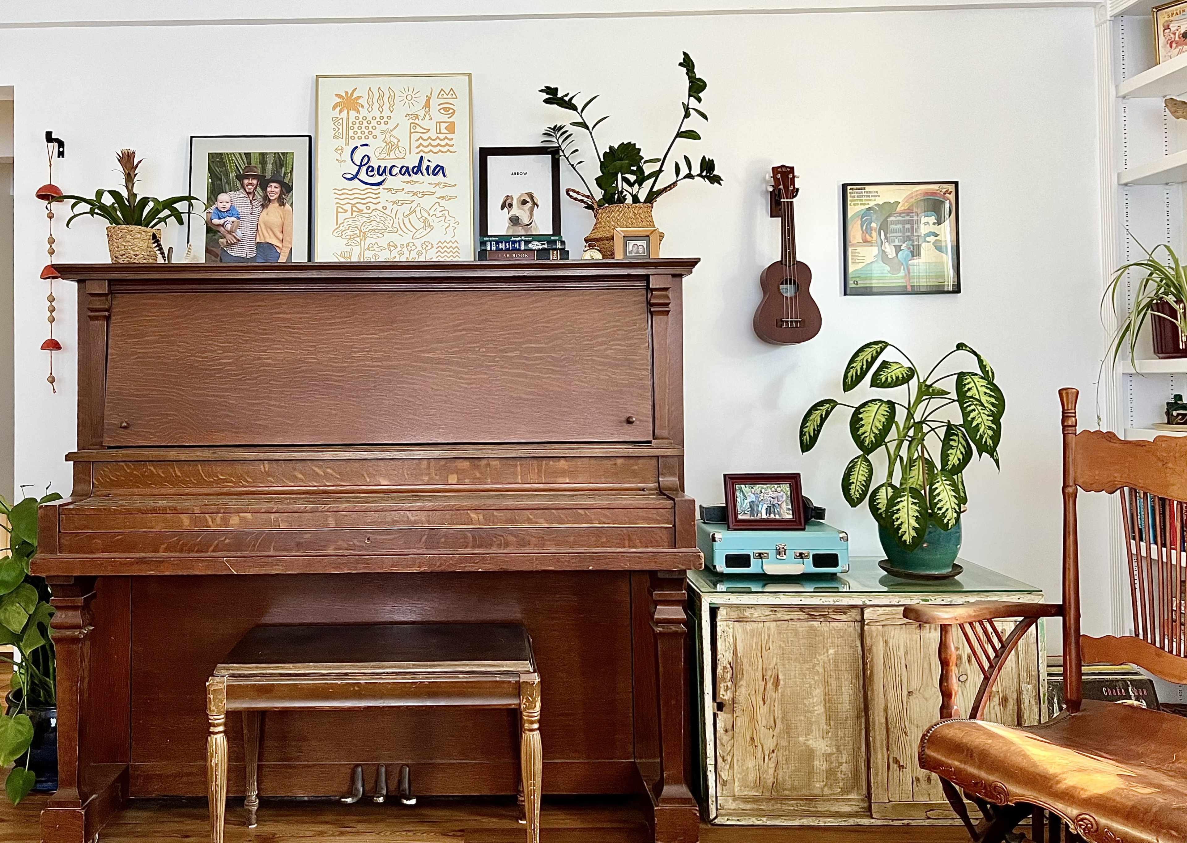 A wooden piano with a decorative vase of plants and framed photos on the wall behind it, alongside a ukulele and a green storage cabinet.