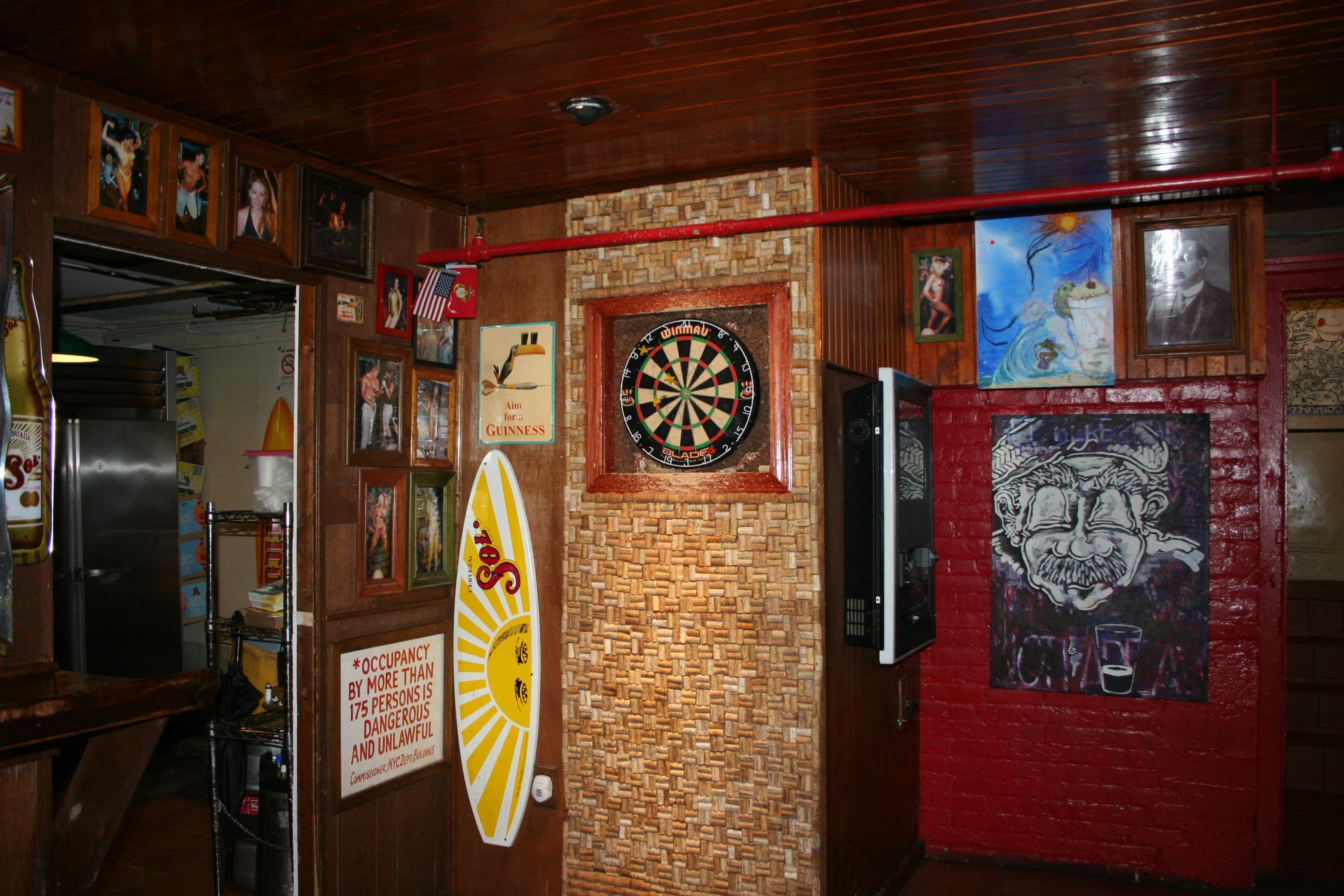 The interior of a bar features wood-paneled walls adorned with framed photos and artwork, a dartboard, and a surfboard leaning against the wall.