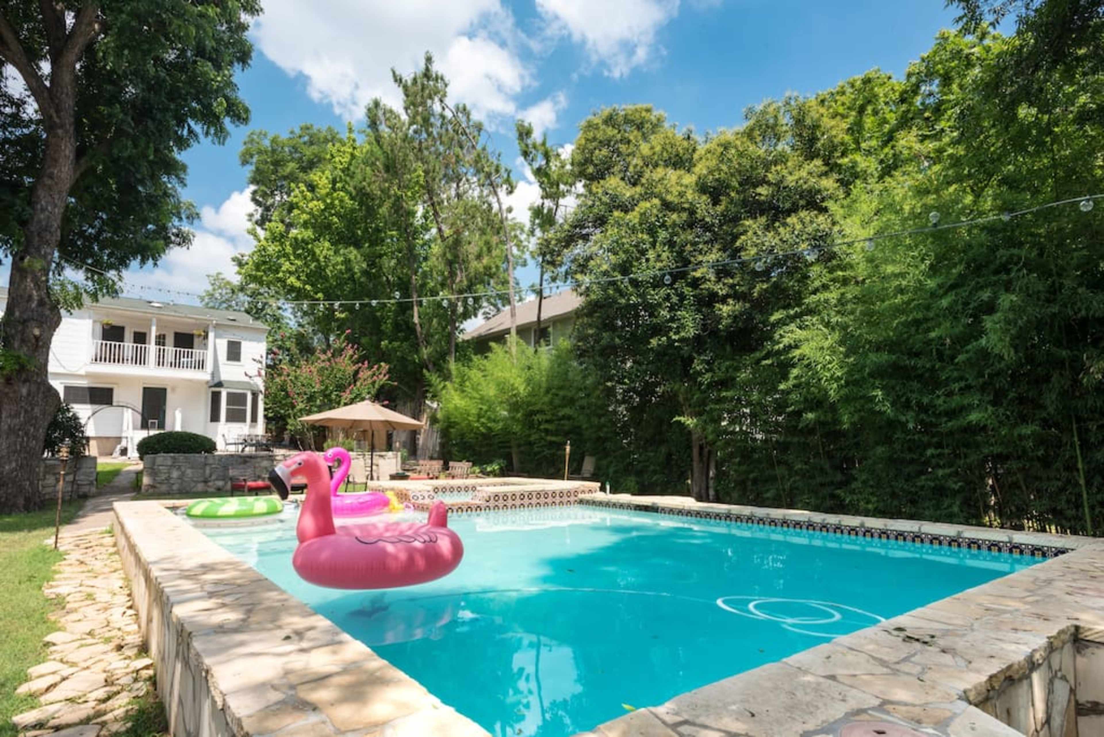 A swimming pool with pink flamingo floaties surrounded by greenery and a two-story house in the background.
