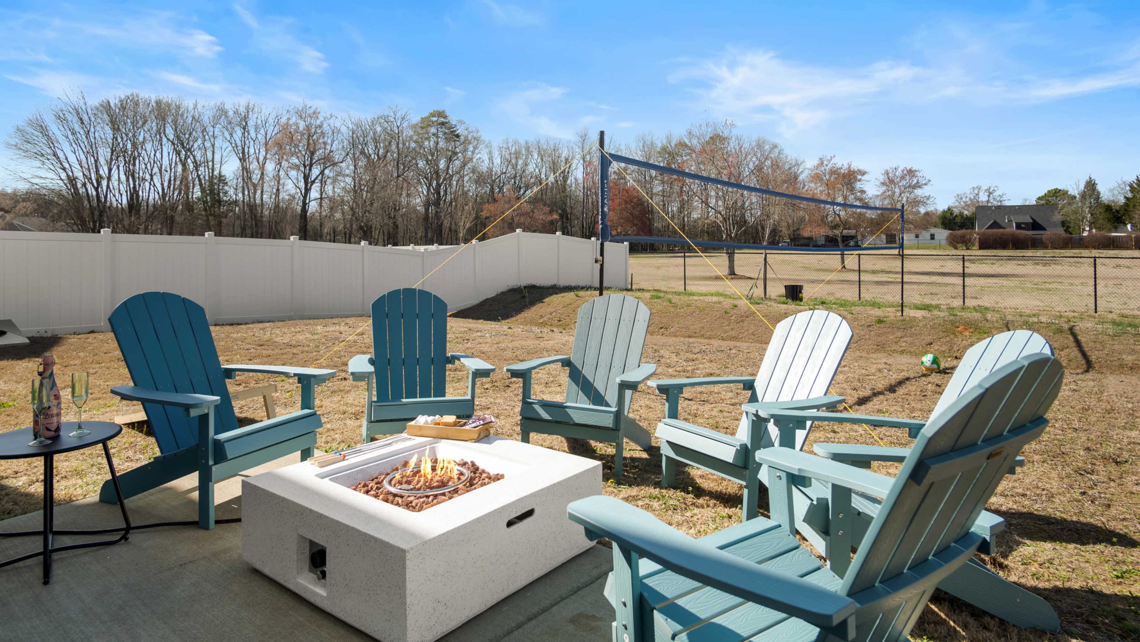 A backyard scene with teal Adirondack chairs arranged around a fire pit, set against a fenced area with a volleyball net in the background.