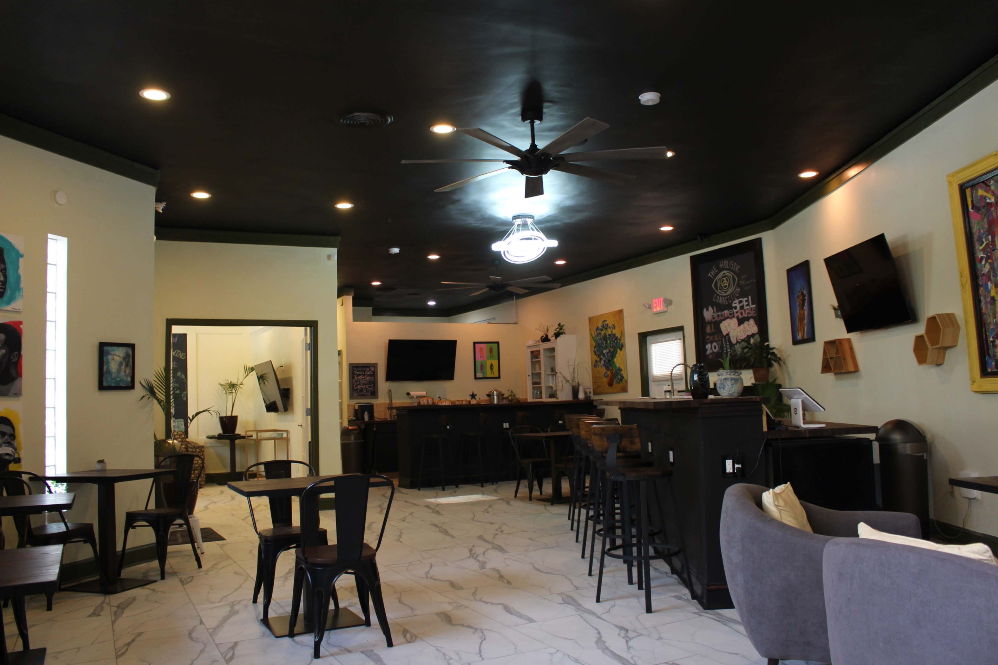 A modern café interior featuring a bar area, seating arrangements, and wall art in a well-lit space with a ceiling fan.
