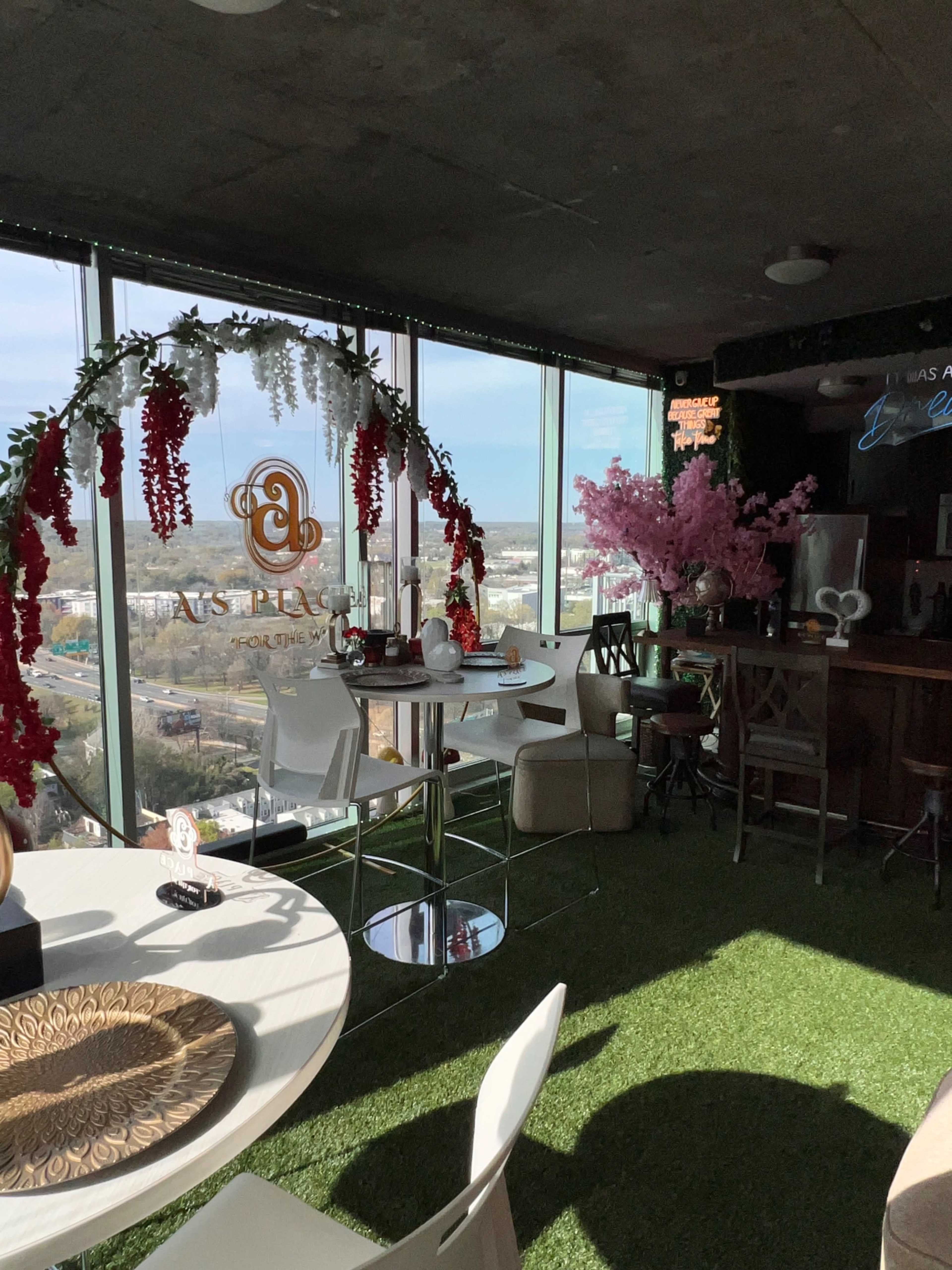 The image shows a modern cafe with large windows offering city views, decorated with artificial flowers and greenery, and featuring tables and chairs arranged for customers.