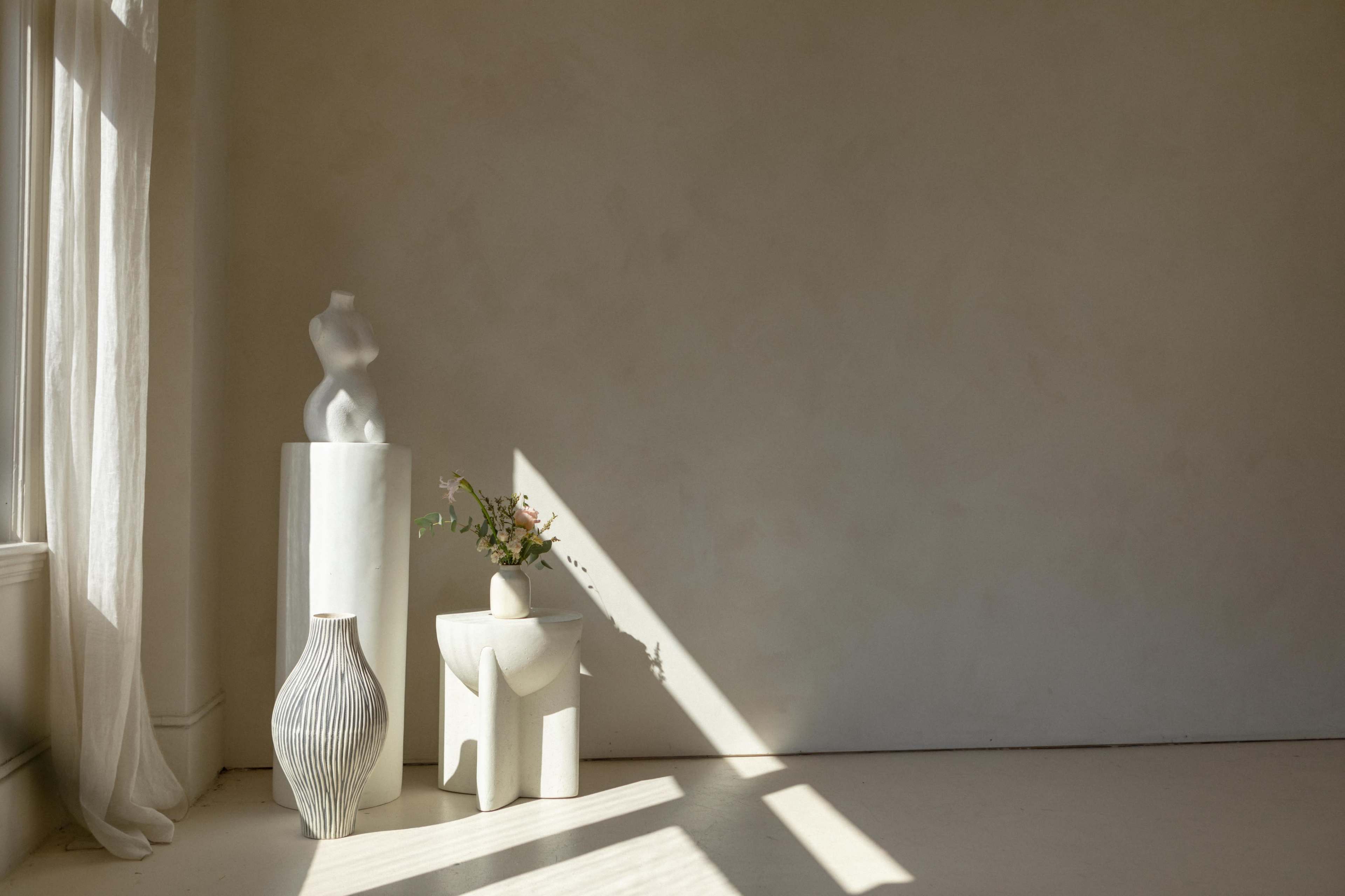A minimalist indoor space with three white decorative pieces, including a sculpture and vases, illuminated by natural light filtering through a window.