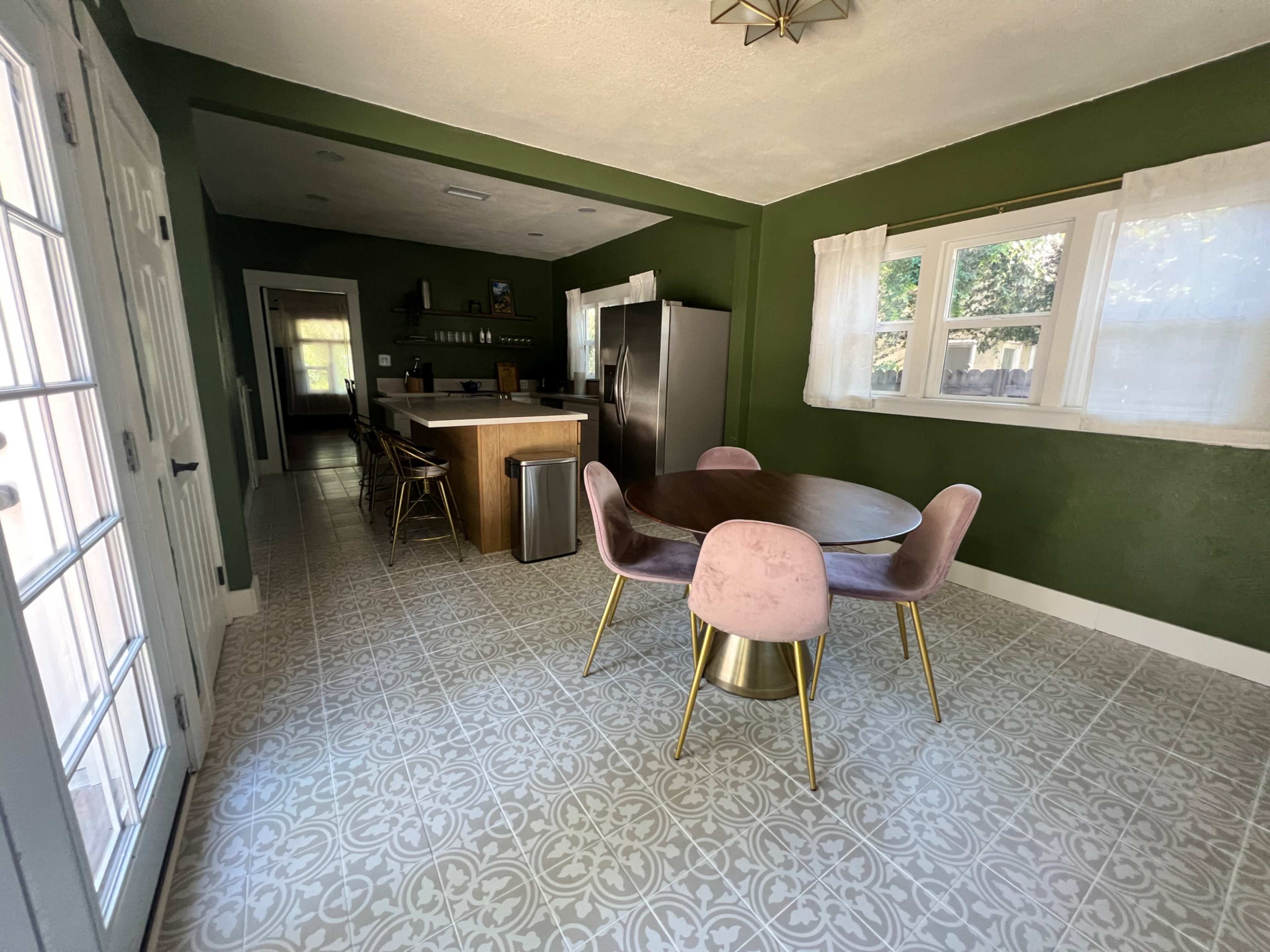 The image shows a modern kitchen with green walls, a circular wooden dining table surrounded by pink chairs, and stainless steel appliances.