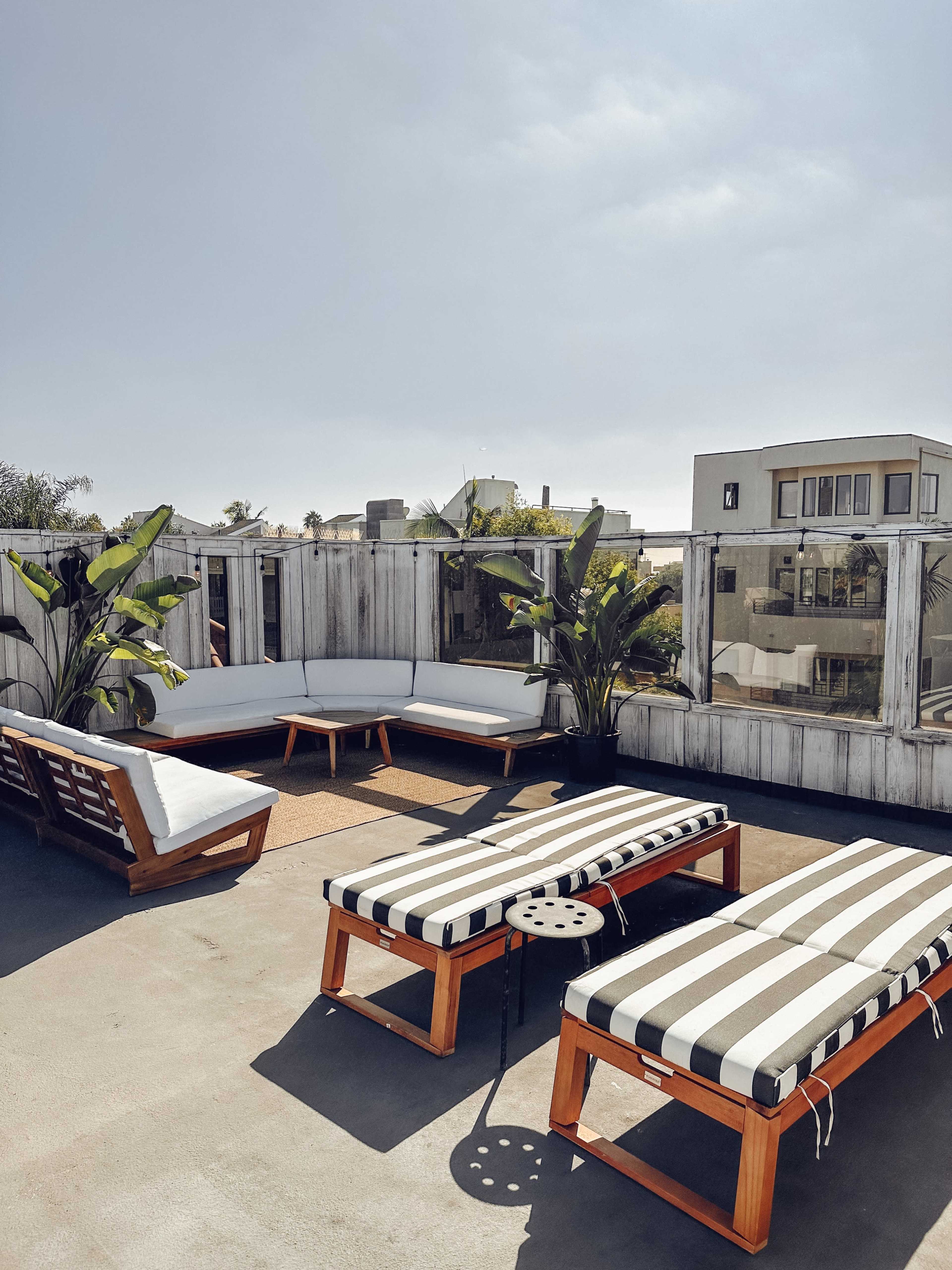 A rooftop terrace features two striped lounge chairs, a wooden coffee table, and a sectional sofa surrounded by potted plants.