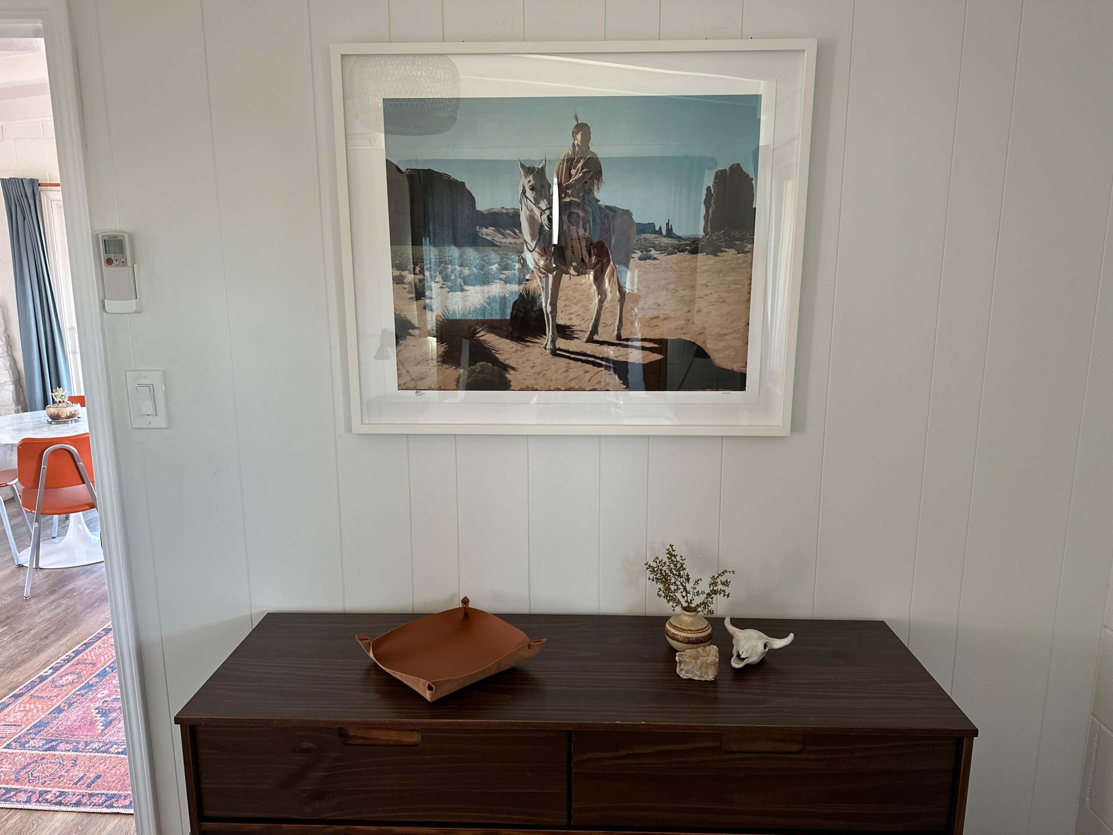 A framed photograph of a person on horseback in a desert landscape is hung on a white-paneled wall above a wooden dresser with decorative items.
