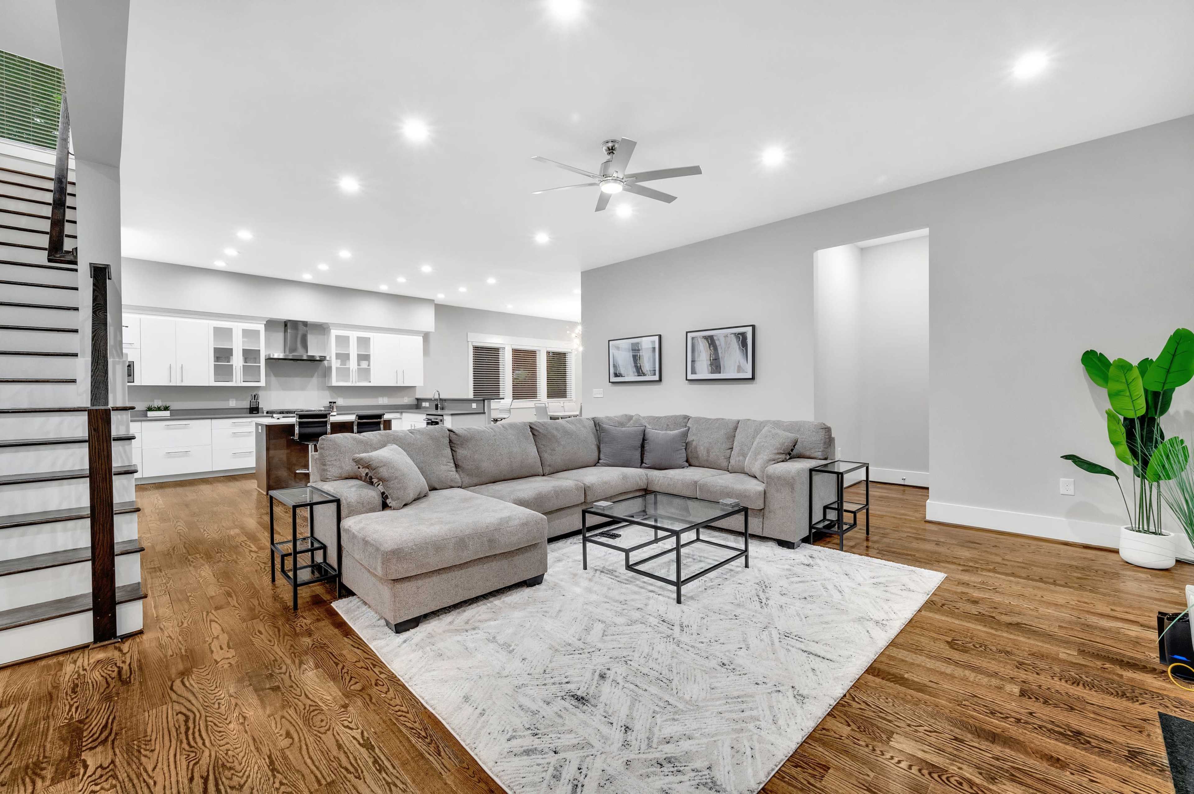 A modern living room featuring a sectional sofa on a patterned rug, a coffee table, and large windows, adjacent to a kitchen with white cabinetry.