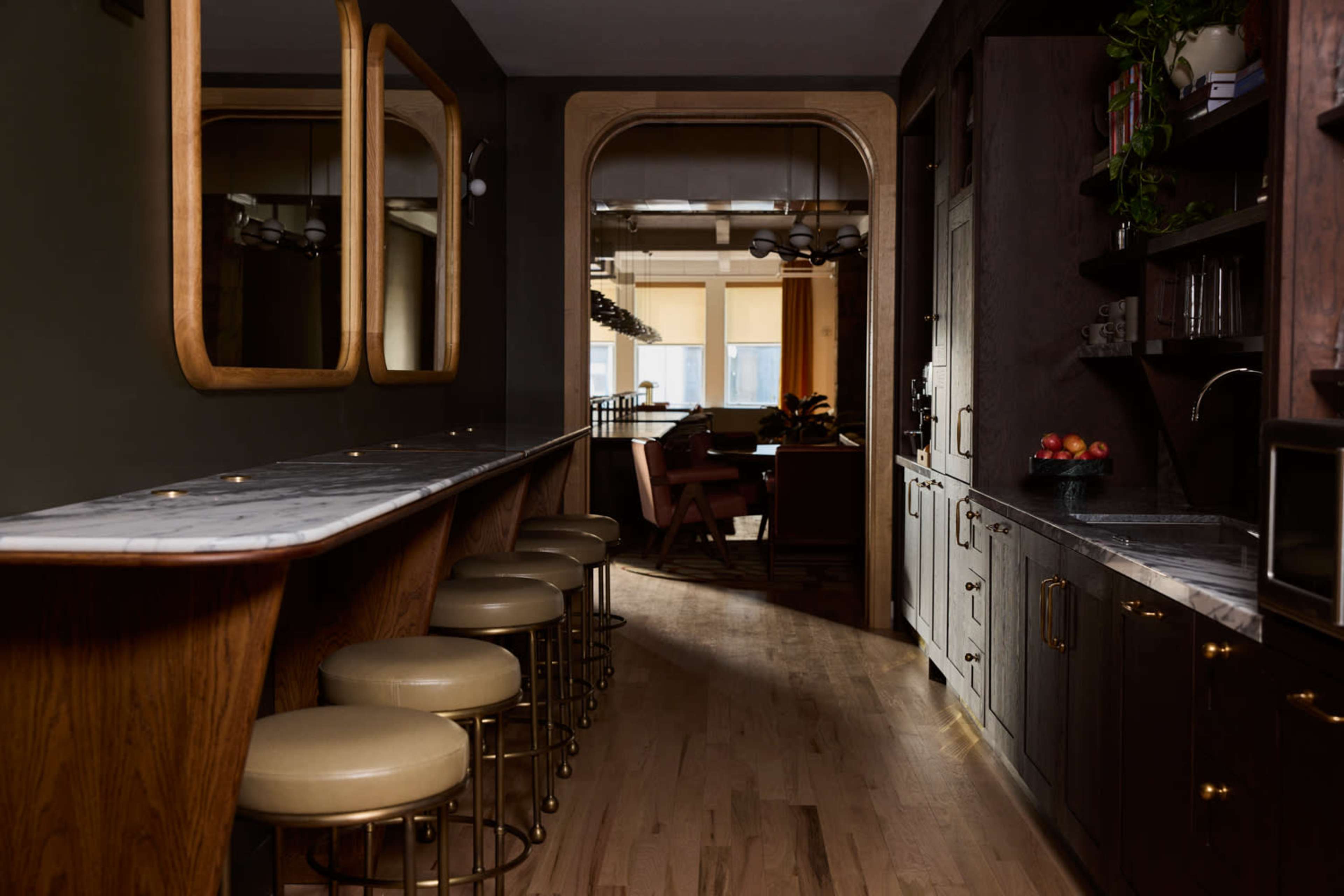The image shows a dimly lit hallway featuring a bar with marble countertops and gold accents, leading to a dining area with large windows and wooden furniture.
