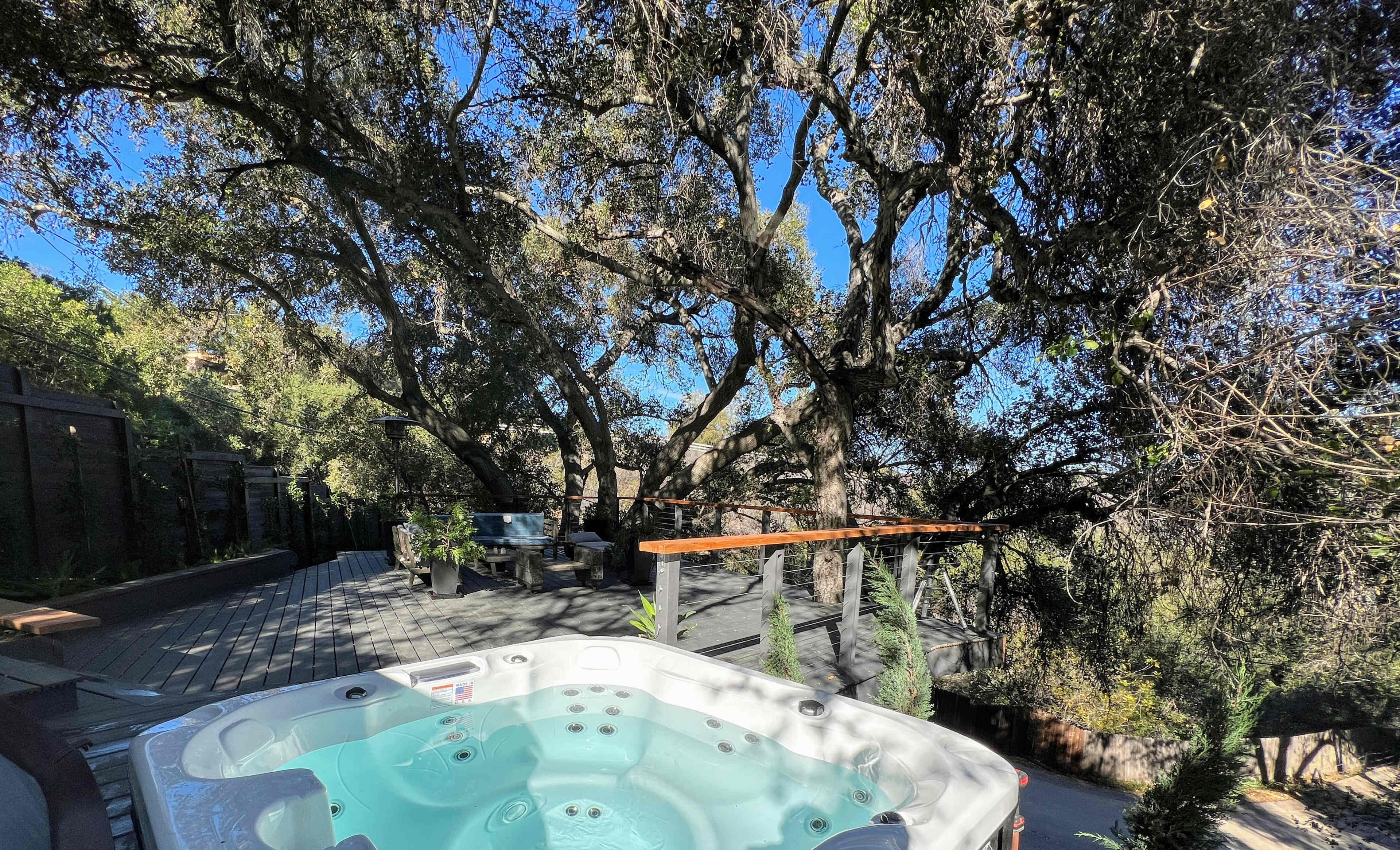 A hot tub is positioned on a deck surrounded by large trees and a shaded seating area.