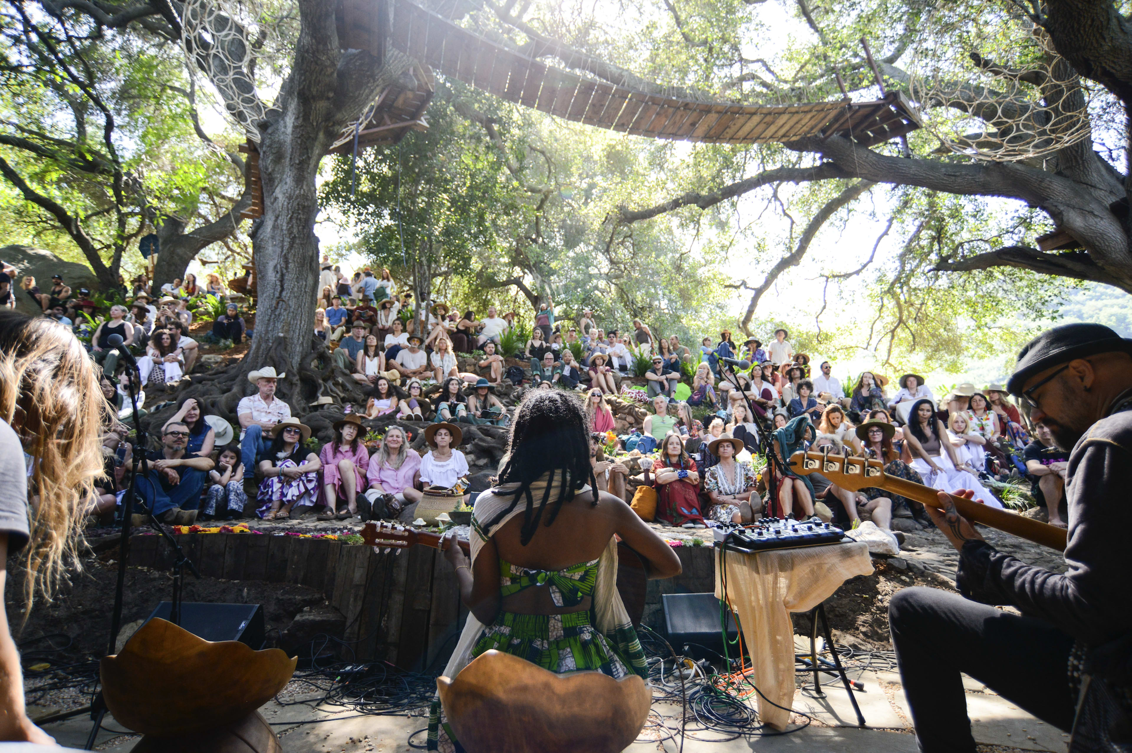 A large audience gathered under trees watches performers on a stage in a natural outdoor setting.