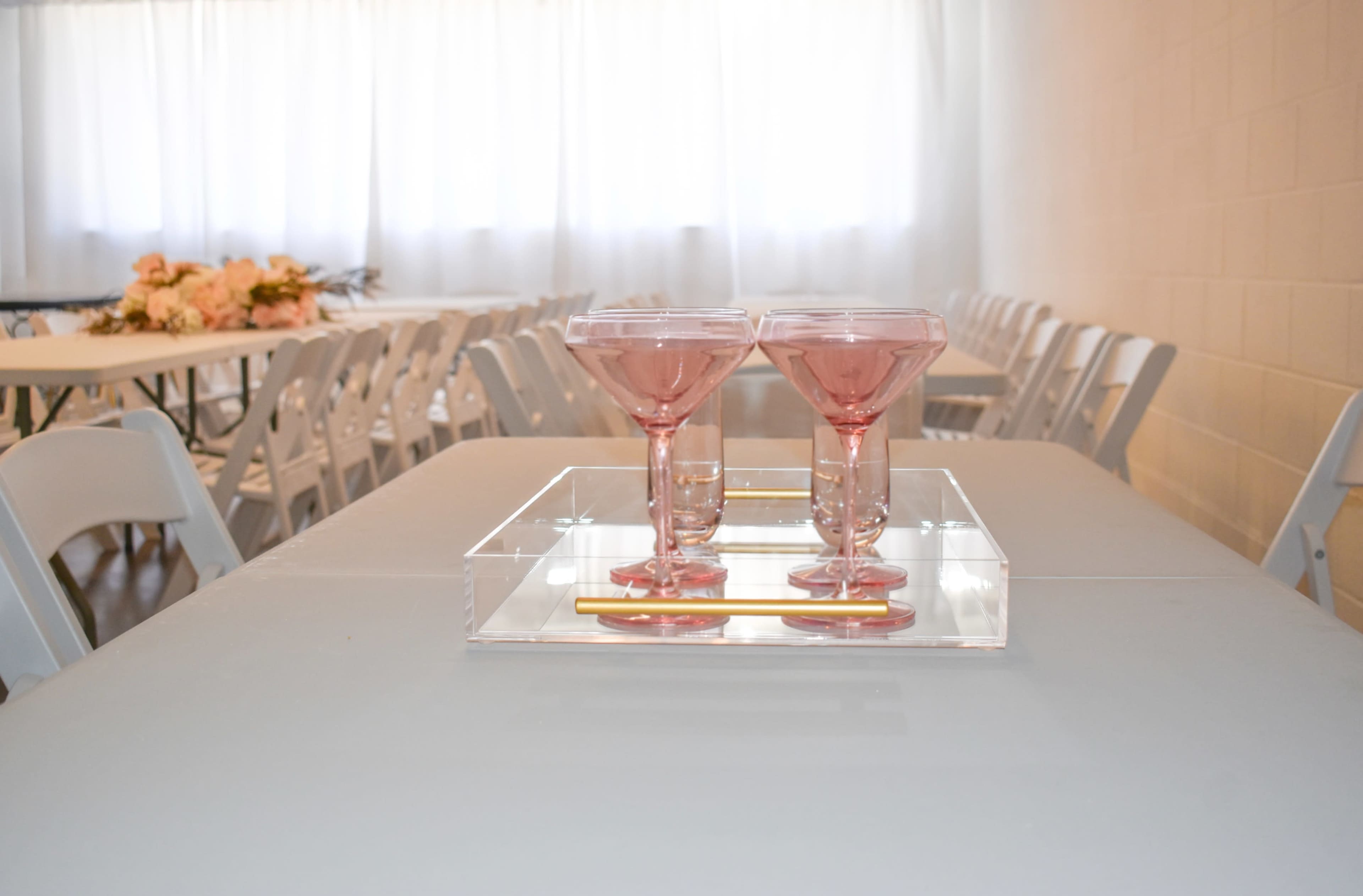 A clear tray holds two pink glasses on a table surrounded by white chairs in a brightly lit room.