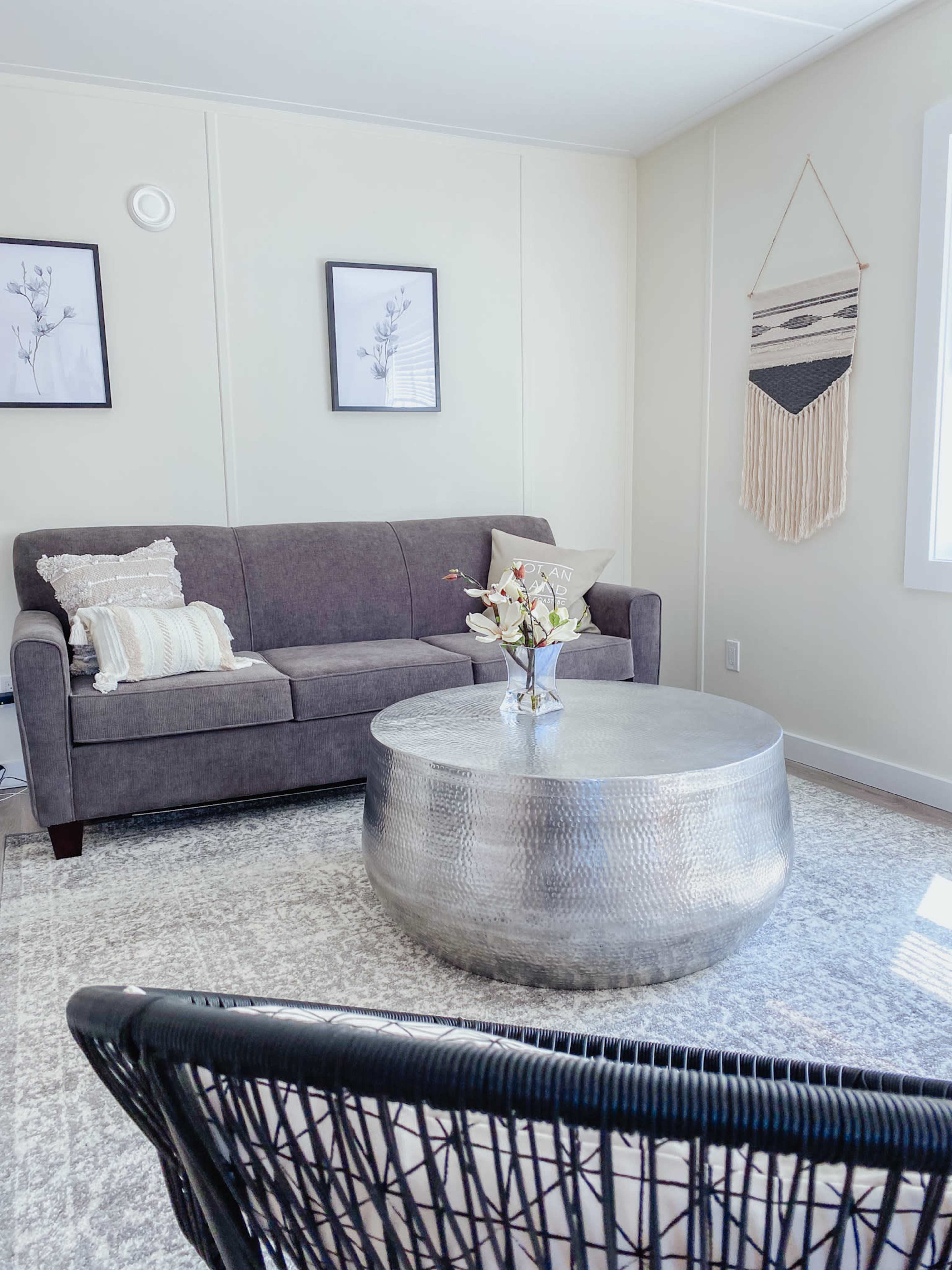 A gray sofa sits next to a round silver coffee table, with a black wicker chair and framed wall art in a light-colored room.