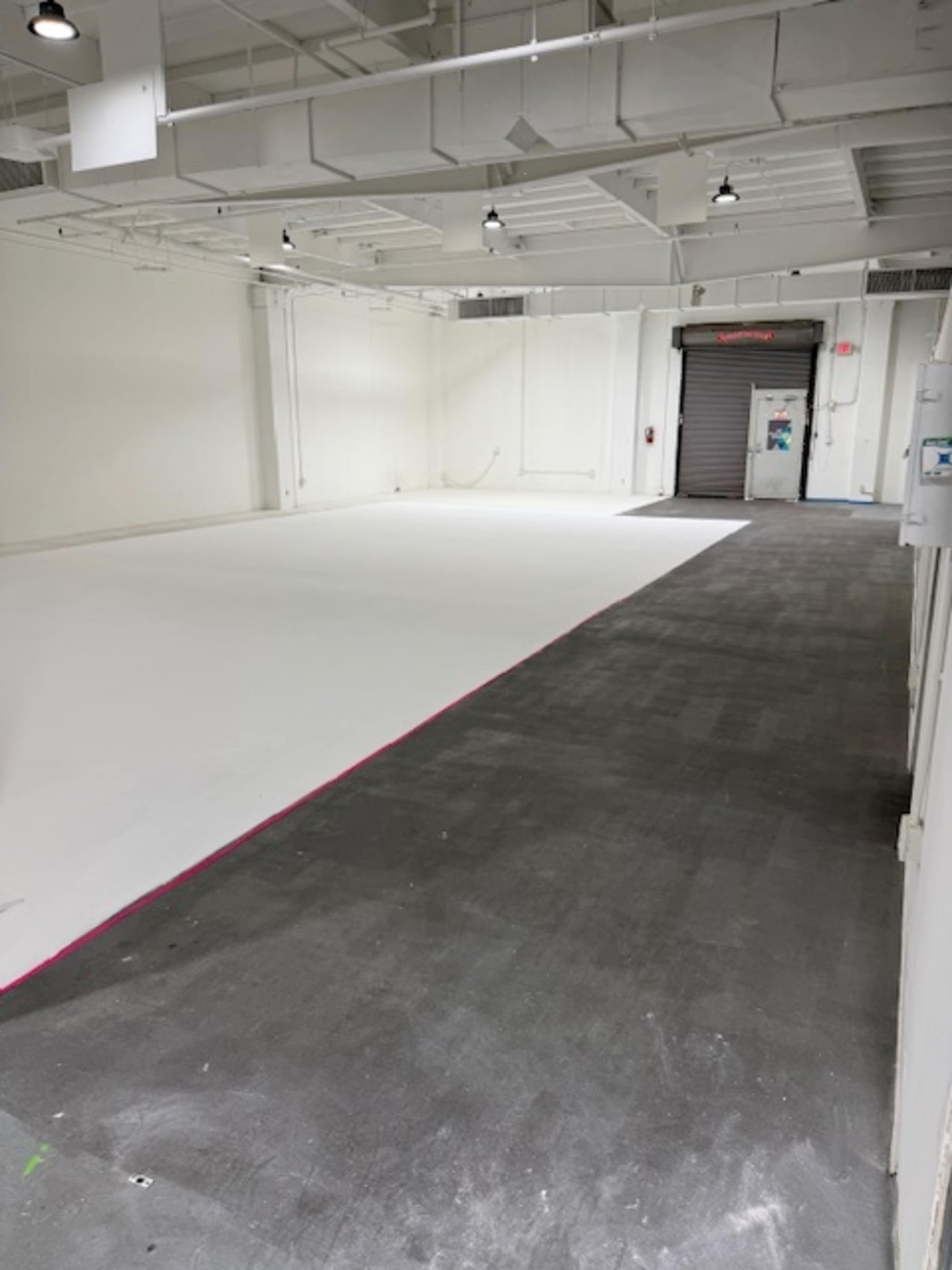 The image shows an empty room with a white floor on one side and a gray floor on the other, leading to a closed garage door.