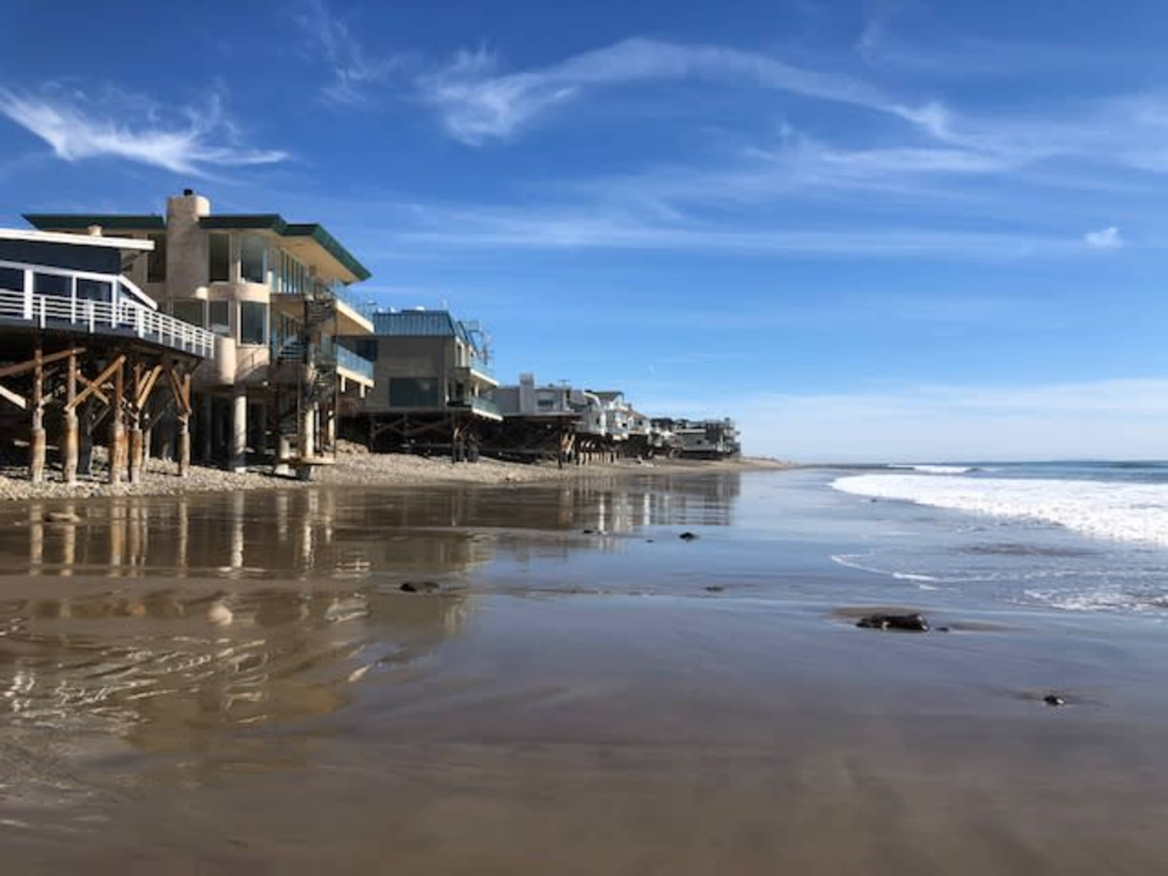 A row of beach houses built on stilts along a sandy shoreline, with gentle waves lapping at the beach.