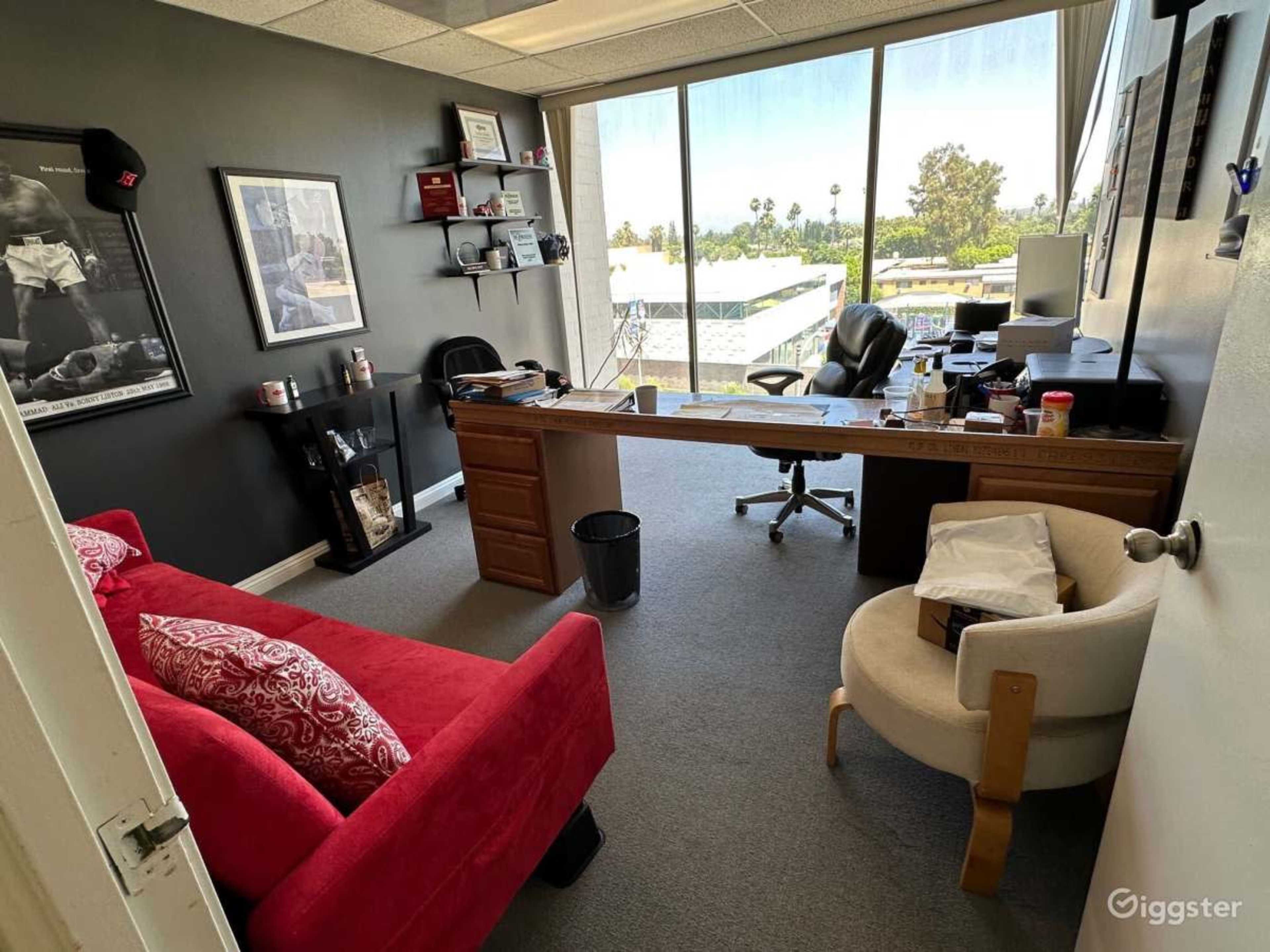 A modern office with a large window, a desk with a chair, and a red sofa next to a white armchair.
