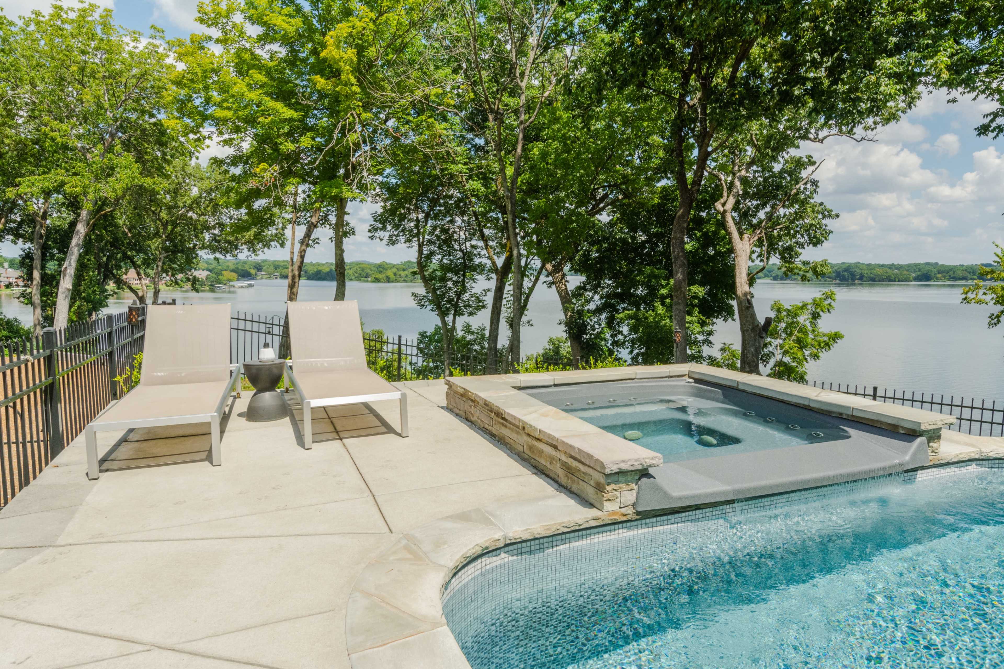 A lakeside patio with two lounge chairs and a hot tub overlooking a serene body of water surrounded by trees.