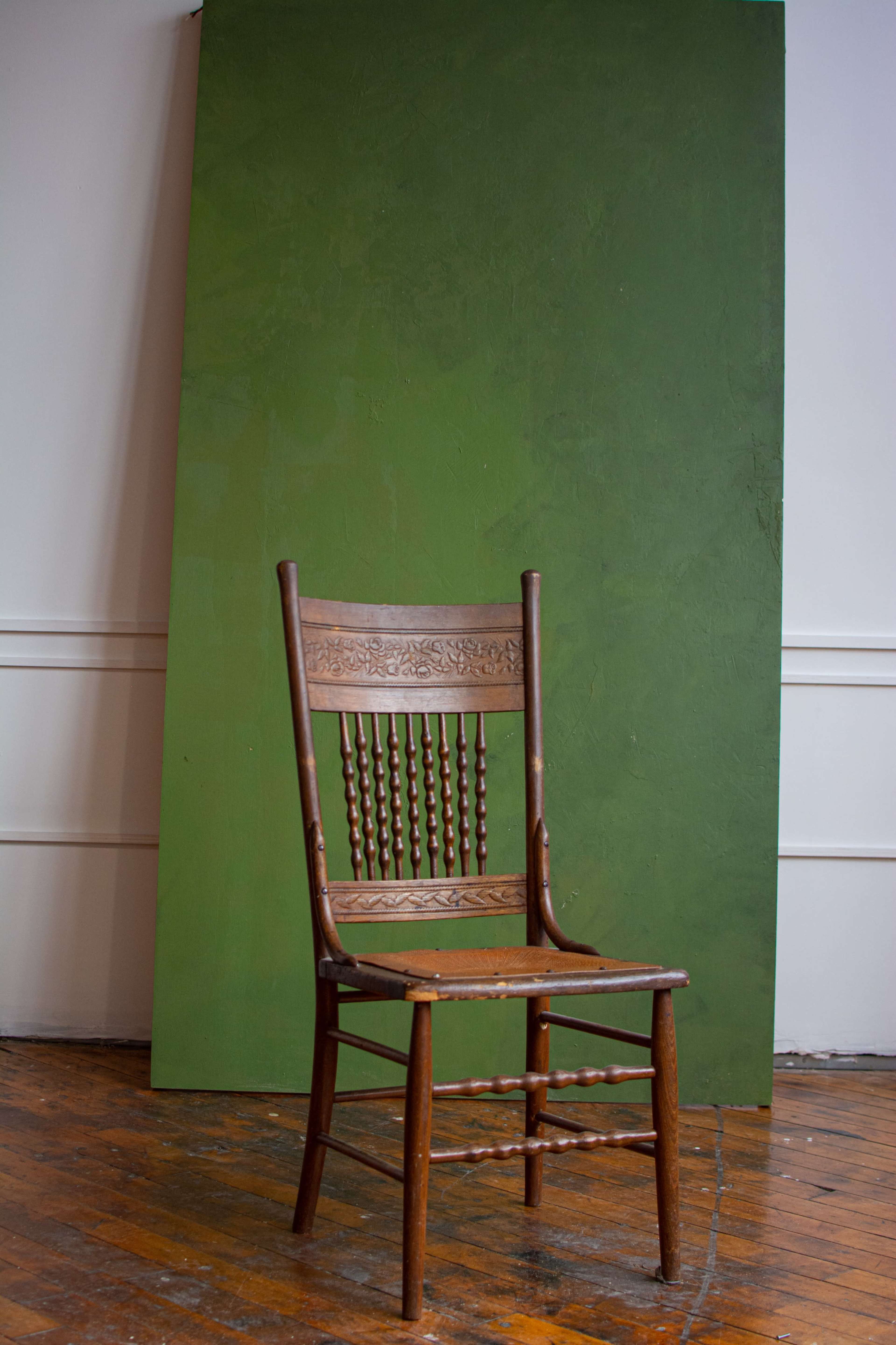 A wooden chair with a decorative backrest stands in front of a large green backdrop on a wooden floor.