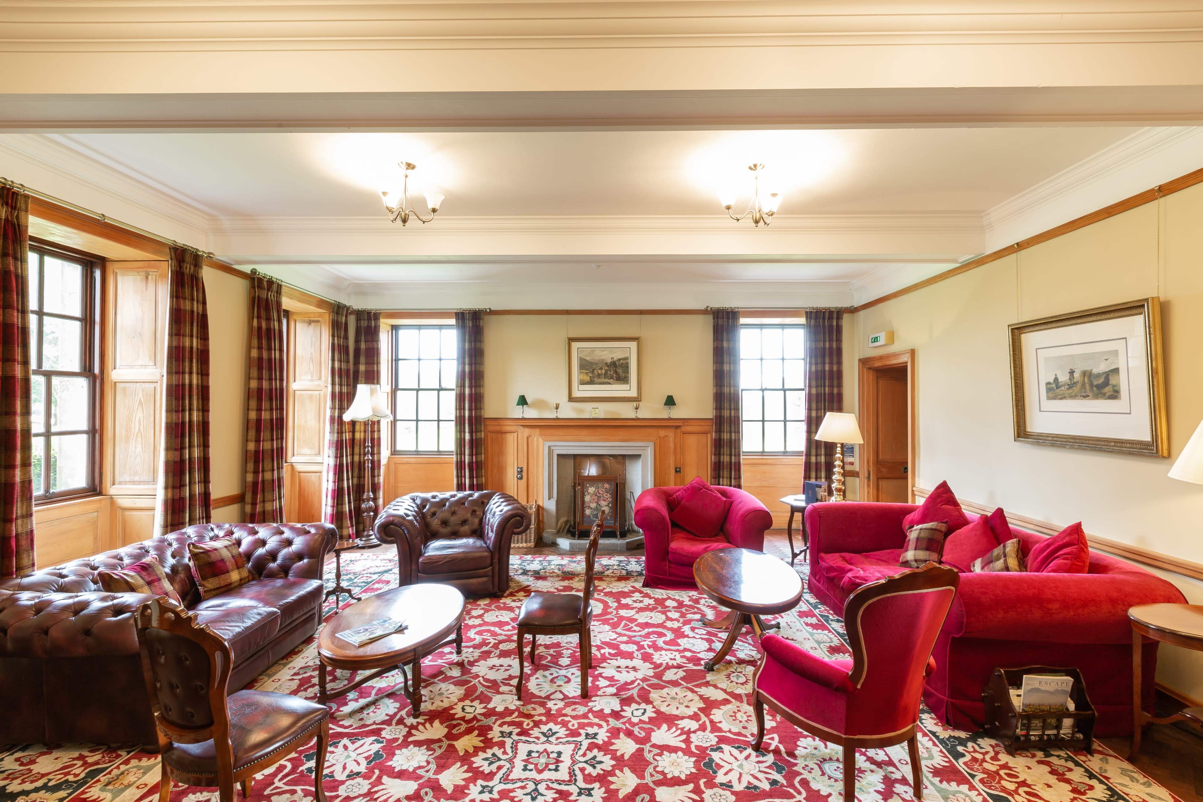 The image shows a spacious, well-lit living room featuring multiple seating options, including leather sofas and red upholstered chairs, alongside a fireplace and decorative rugs.