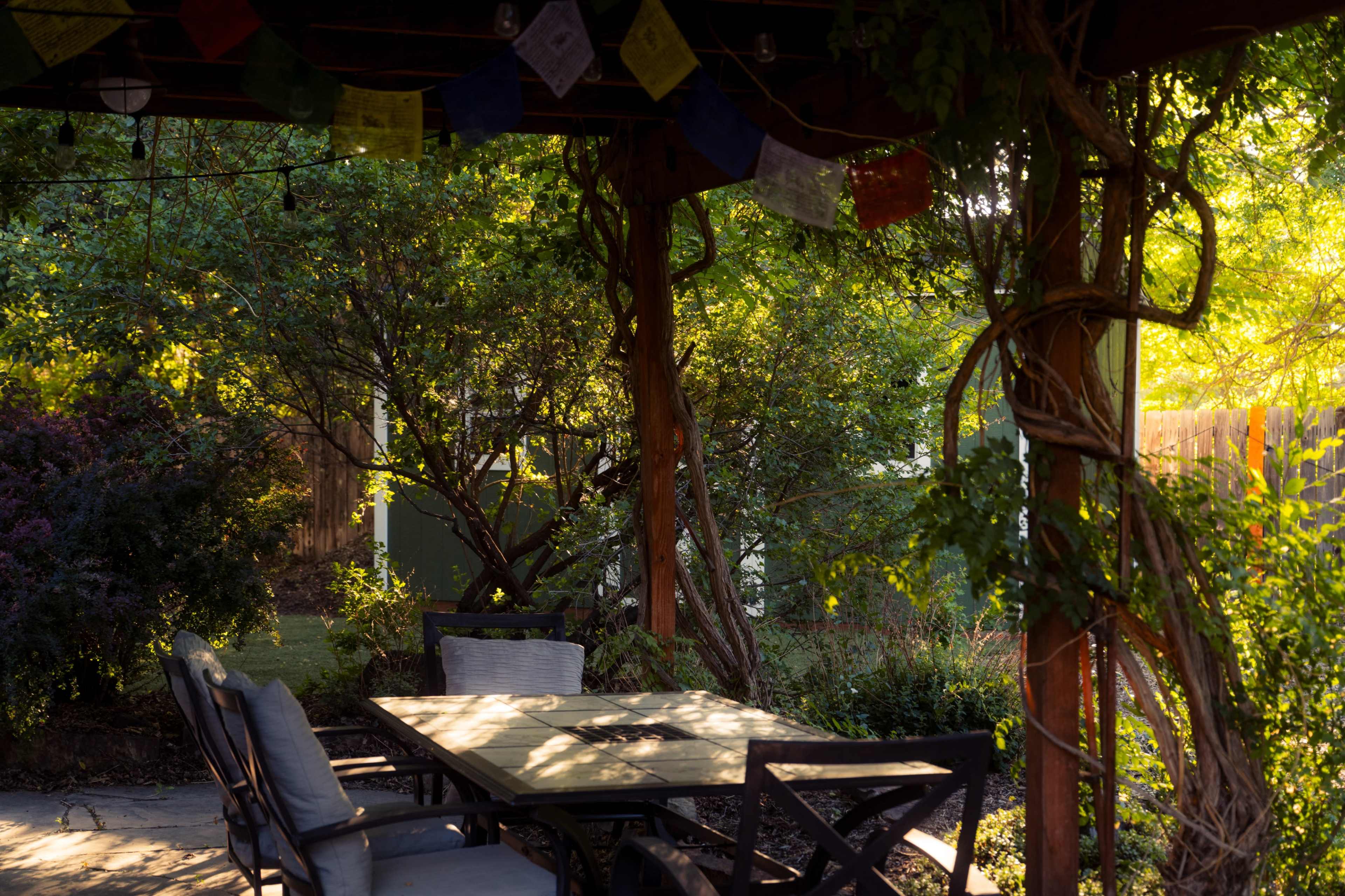 A wooden pergola is adorned with colorful prayer flags and surrounded by lush greenery, featuring a table and chairs underneath.