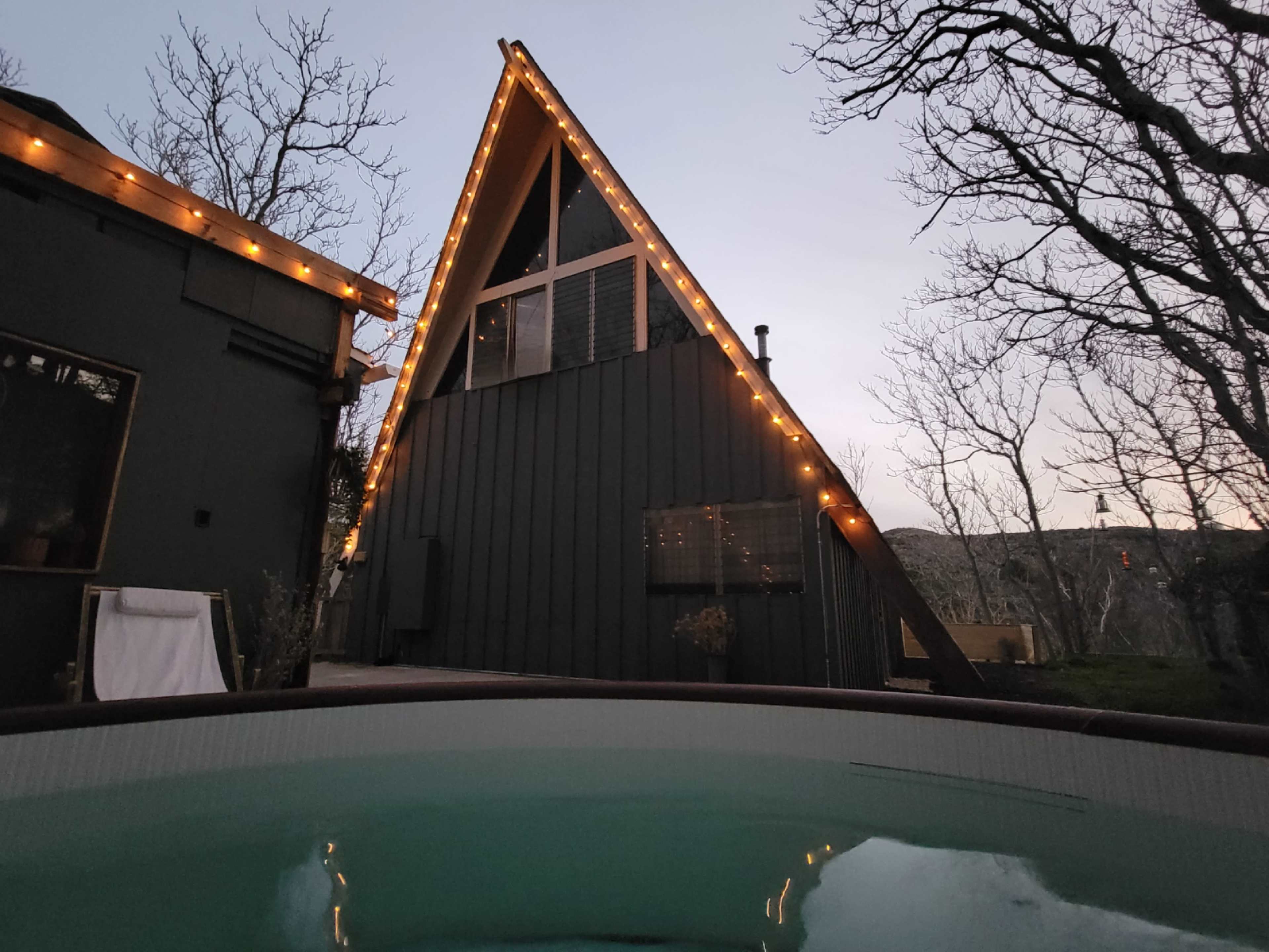 An A-frame cabin with string lights is positioned near a hot tub, surrounded by bare trees at dusk.