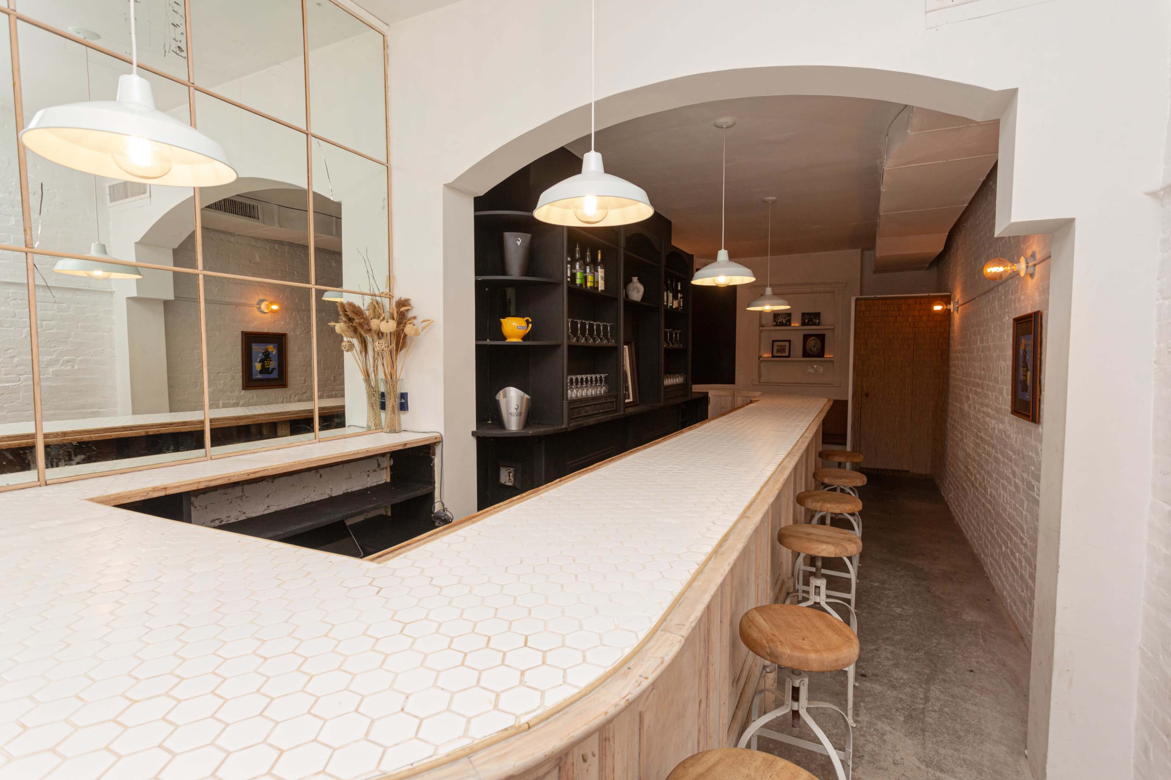 The image shows a modern bar setup with a white hexagonal tiled counter, wooden stools, and illuminated shelving in a softly lit space.