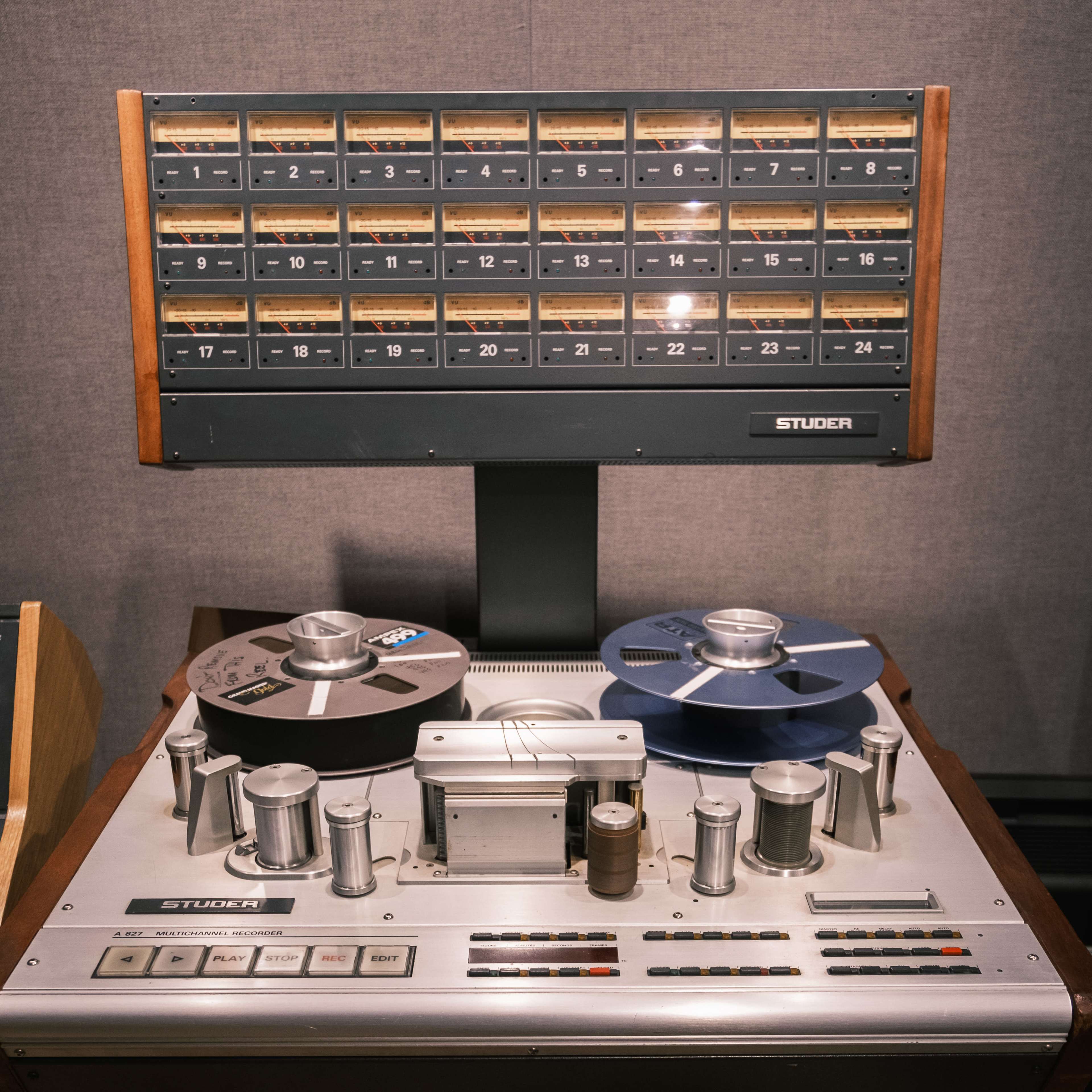 The image shows a vintage Studer tape machine with a large control panel and multiple tape reels, positioned in front of a display board featuring labeled audio tape slots.