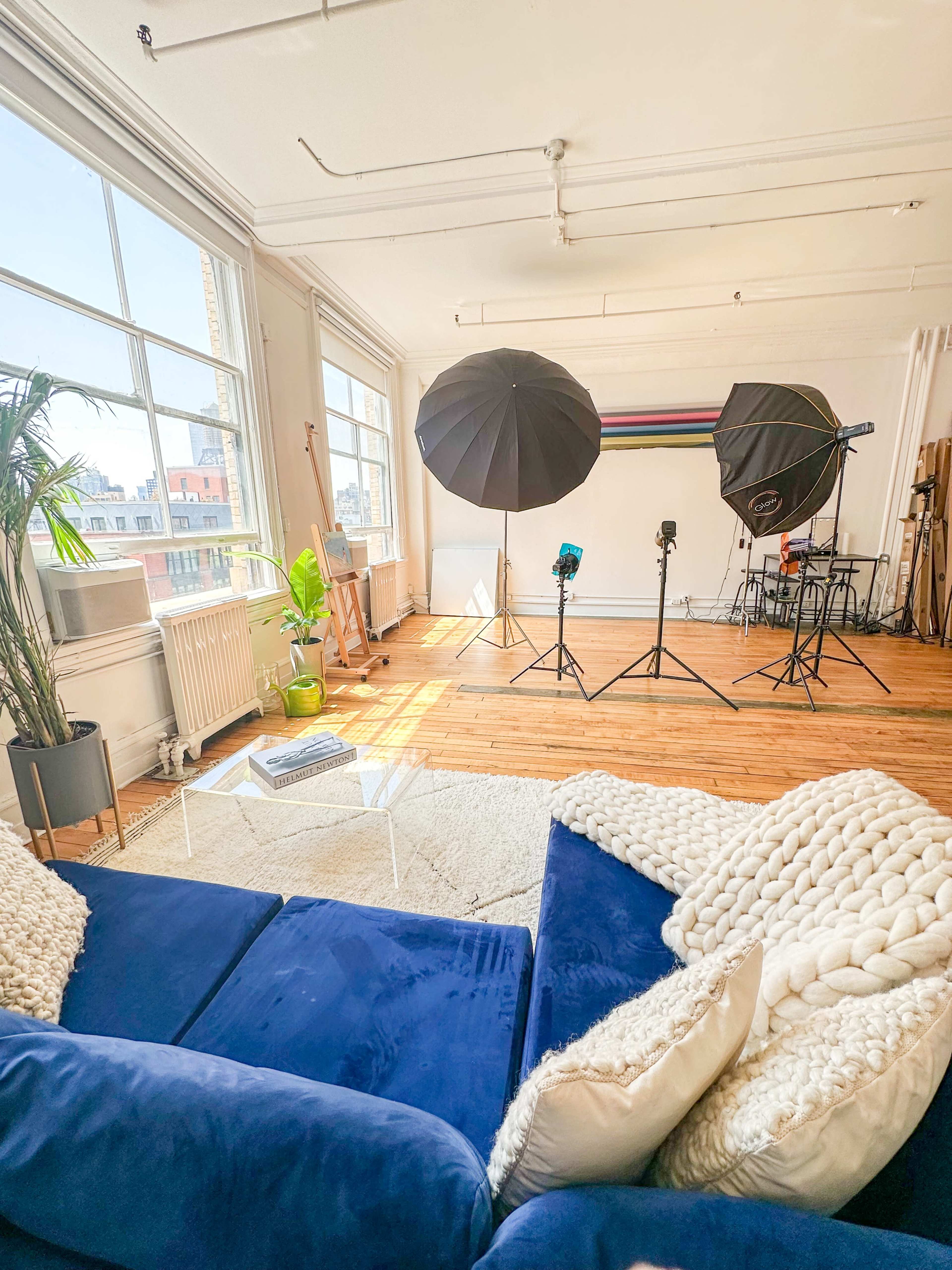 A spacious studio with large windows, featuring a blue sofa, a rug, and two photography umbrellas set up for a shoot.