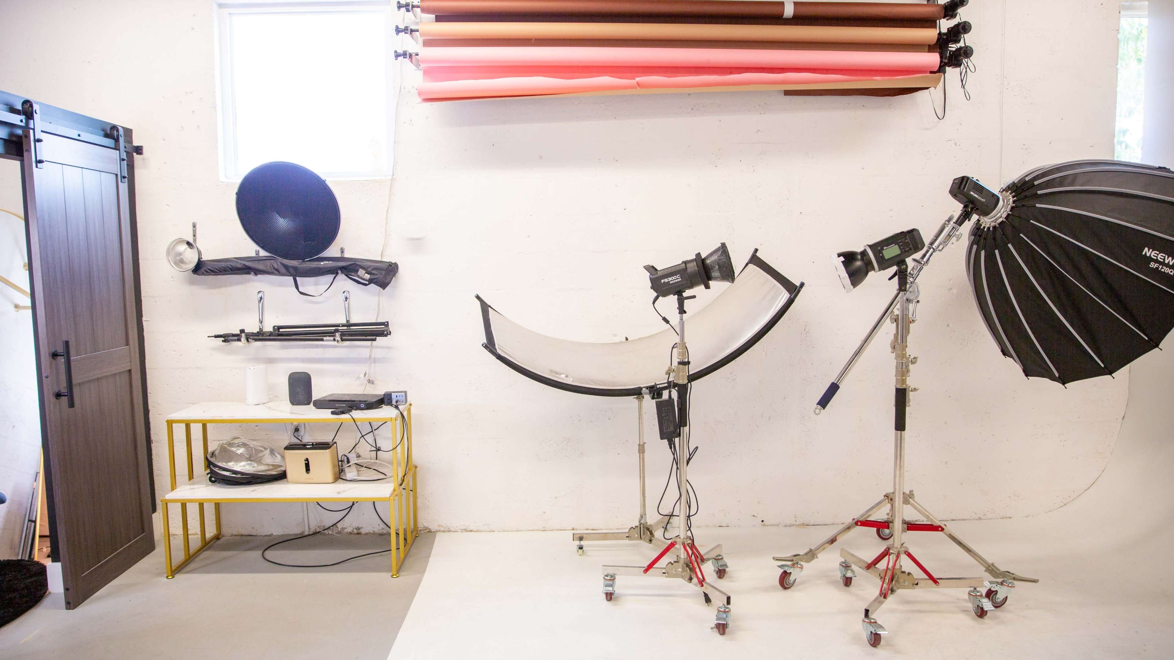 The image shows a photography studio setup with various lighting equipment, a backdrop, a round table, and a wooden door along one wall.