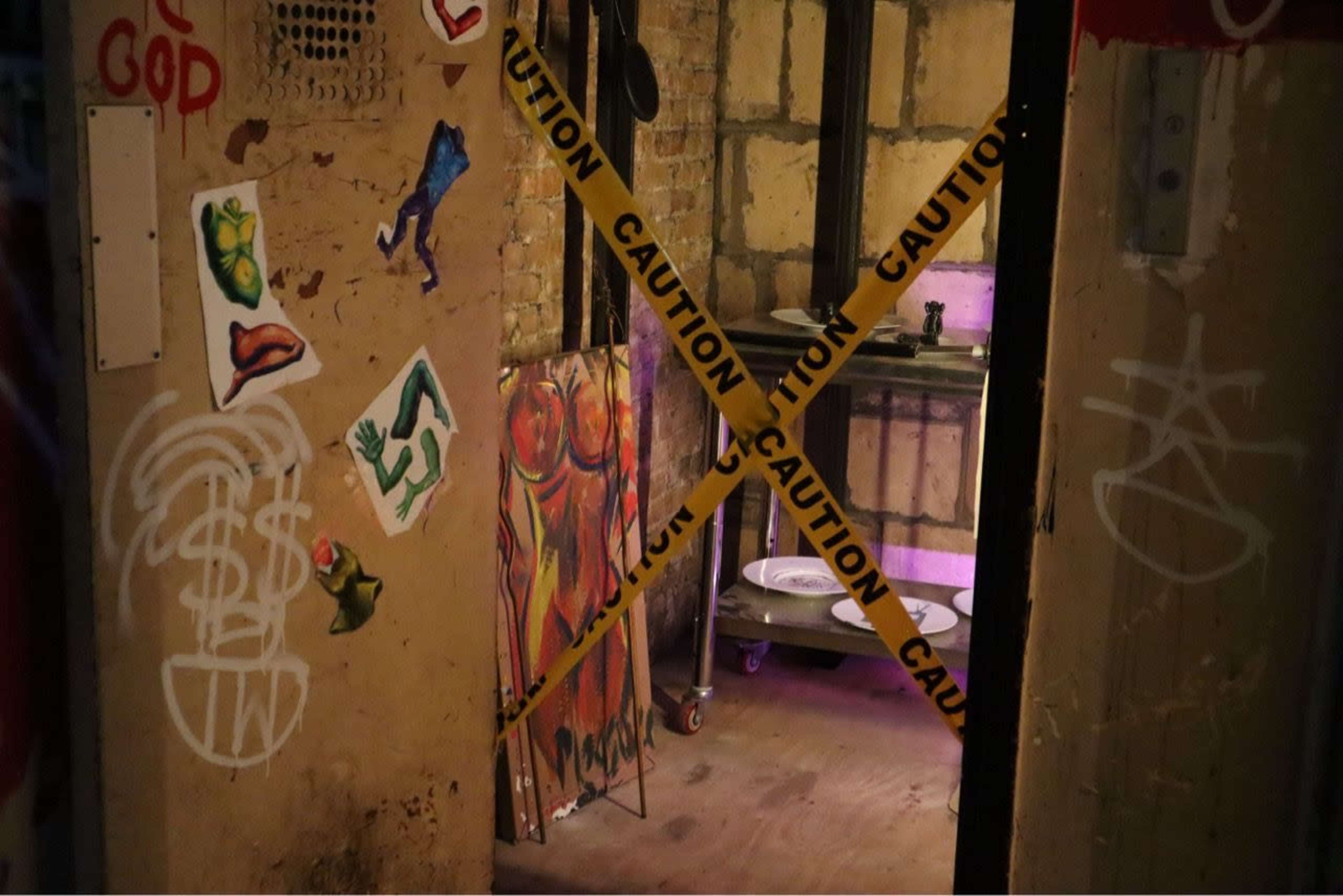 An open doorway with "Caution" tape across it leads into a dimly lit room featuring walls adorned with various colorful stickers and graffiti, alongside a cart holding kitchenware.