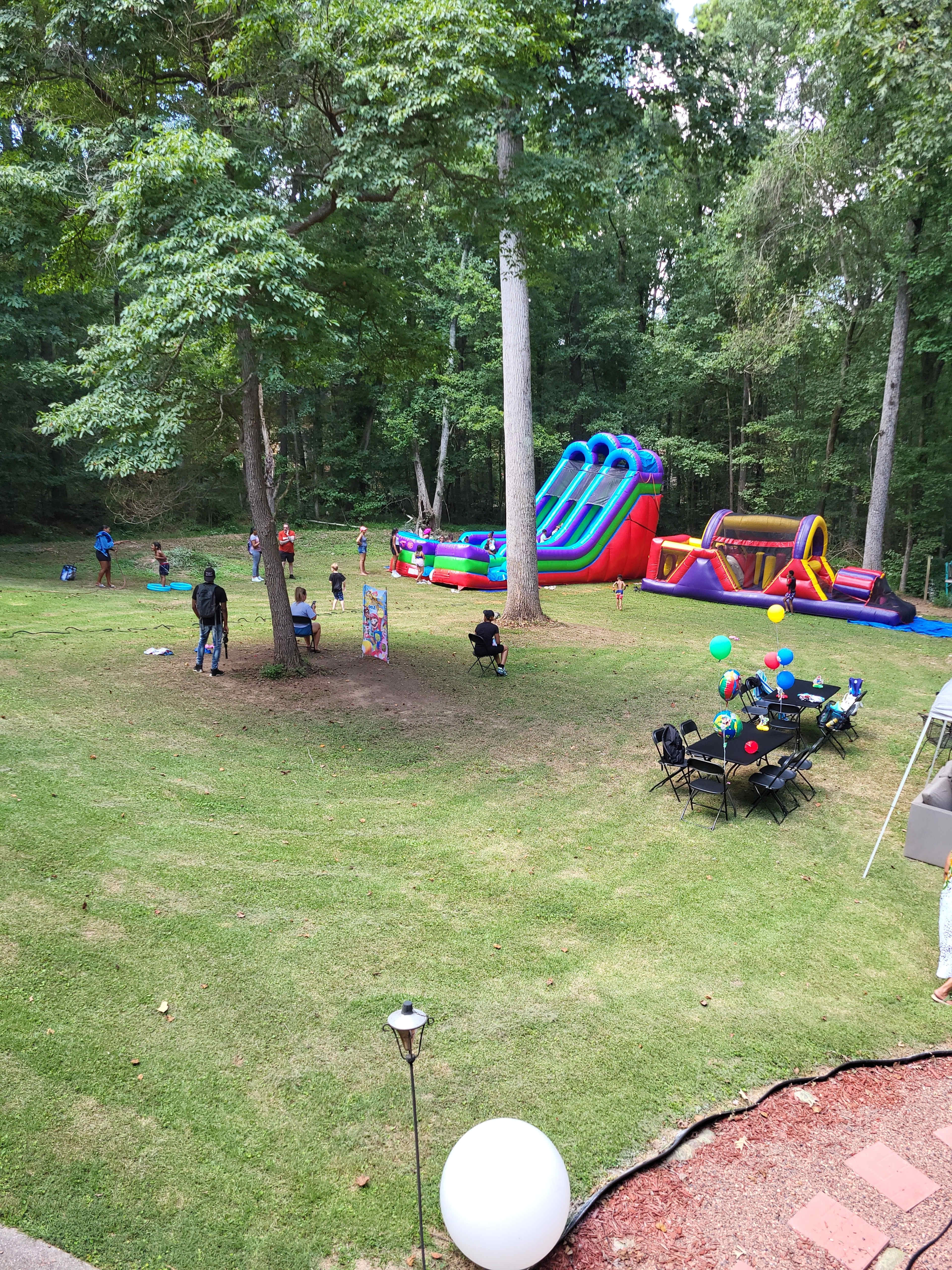 The image shows a backyard with inflatable slides and games, children playing, and tables set with balloons and chairs.