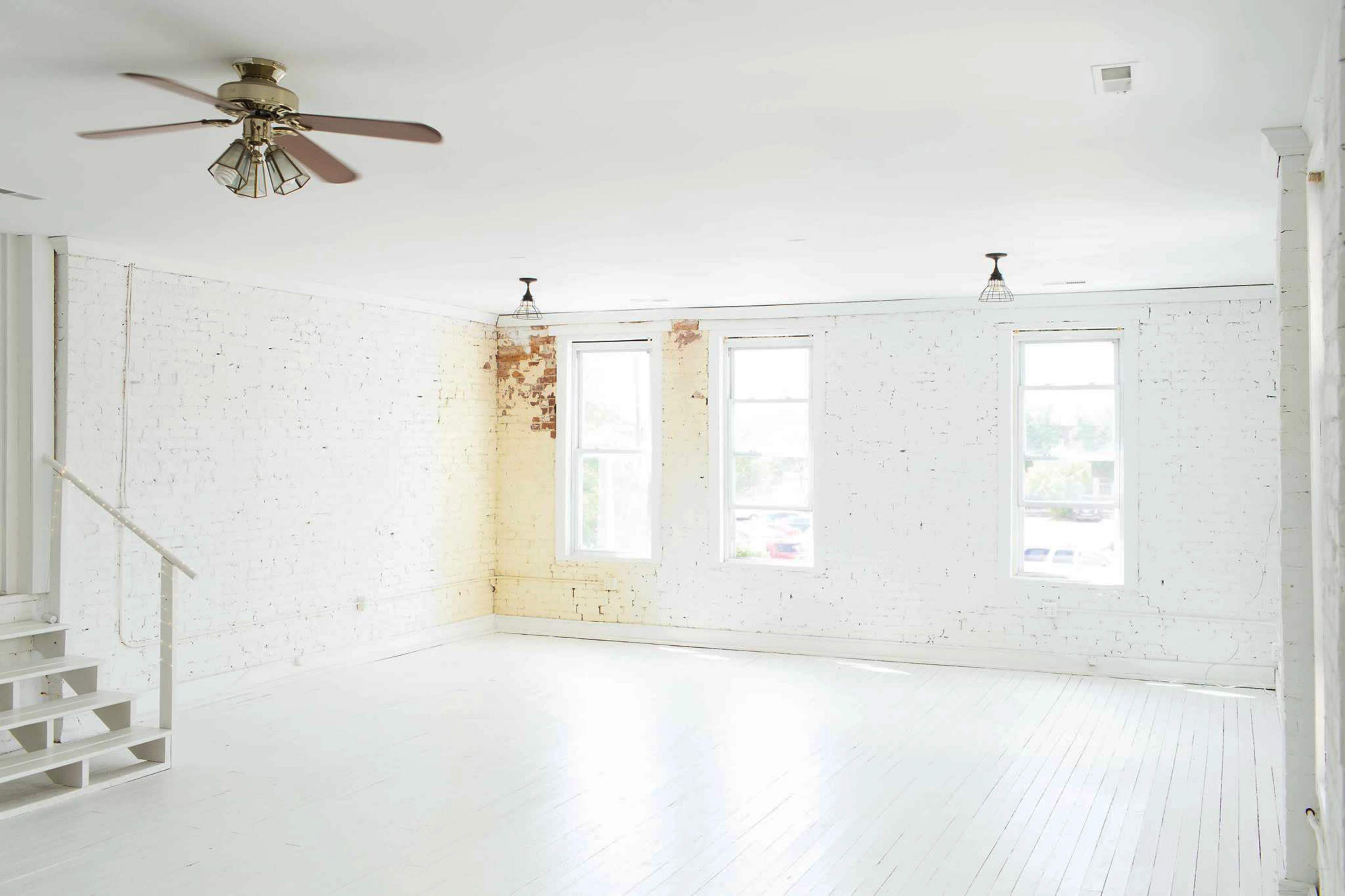 A spacious, empty room features white brick walls, large windows, and a ceiling fan.