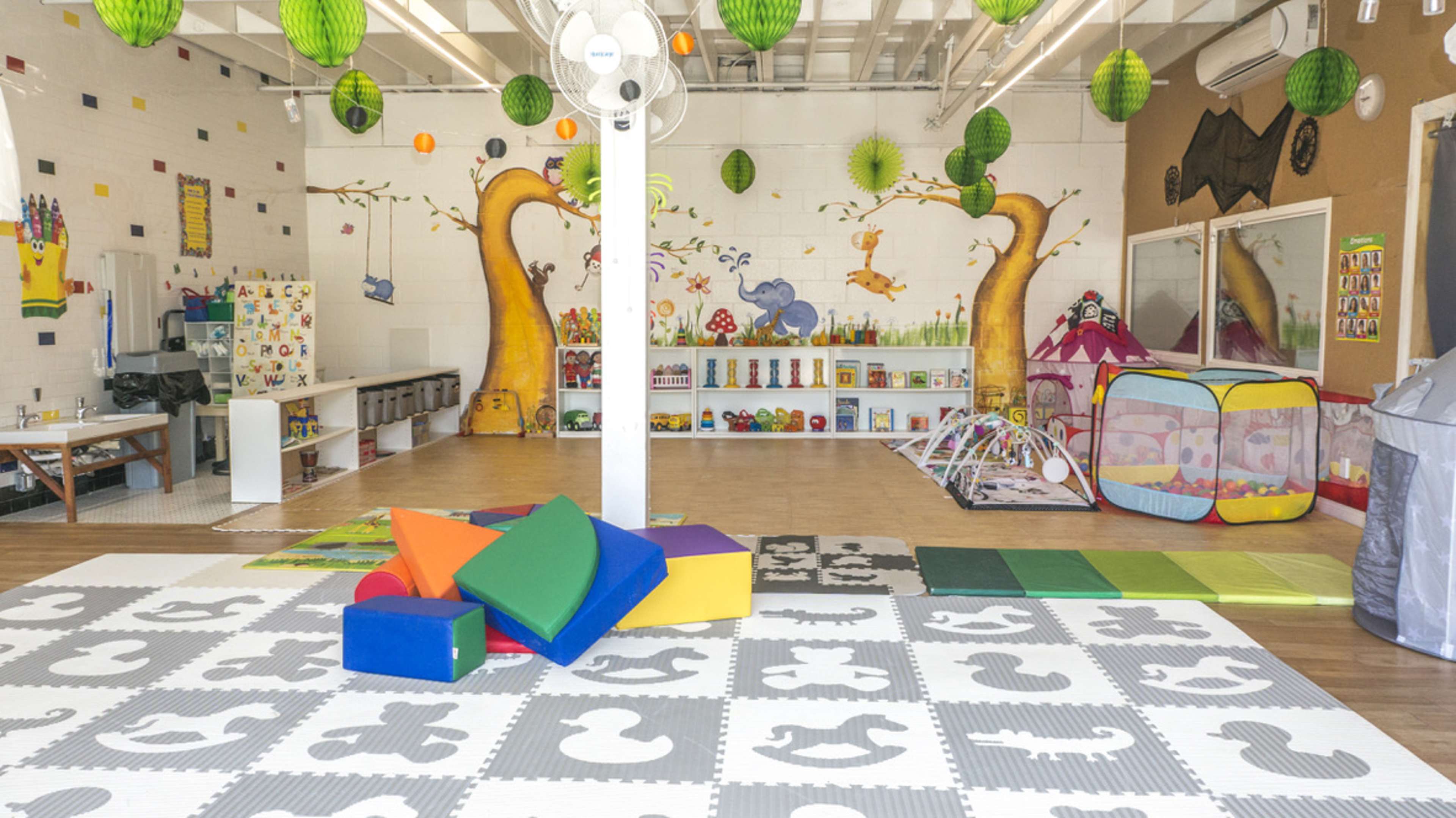 A brightly colored playroom filled with toys, soft mats, and a play tent, featuring murals on the walls and green decorative lanterns overhead.